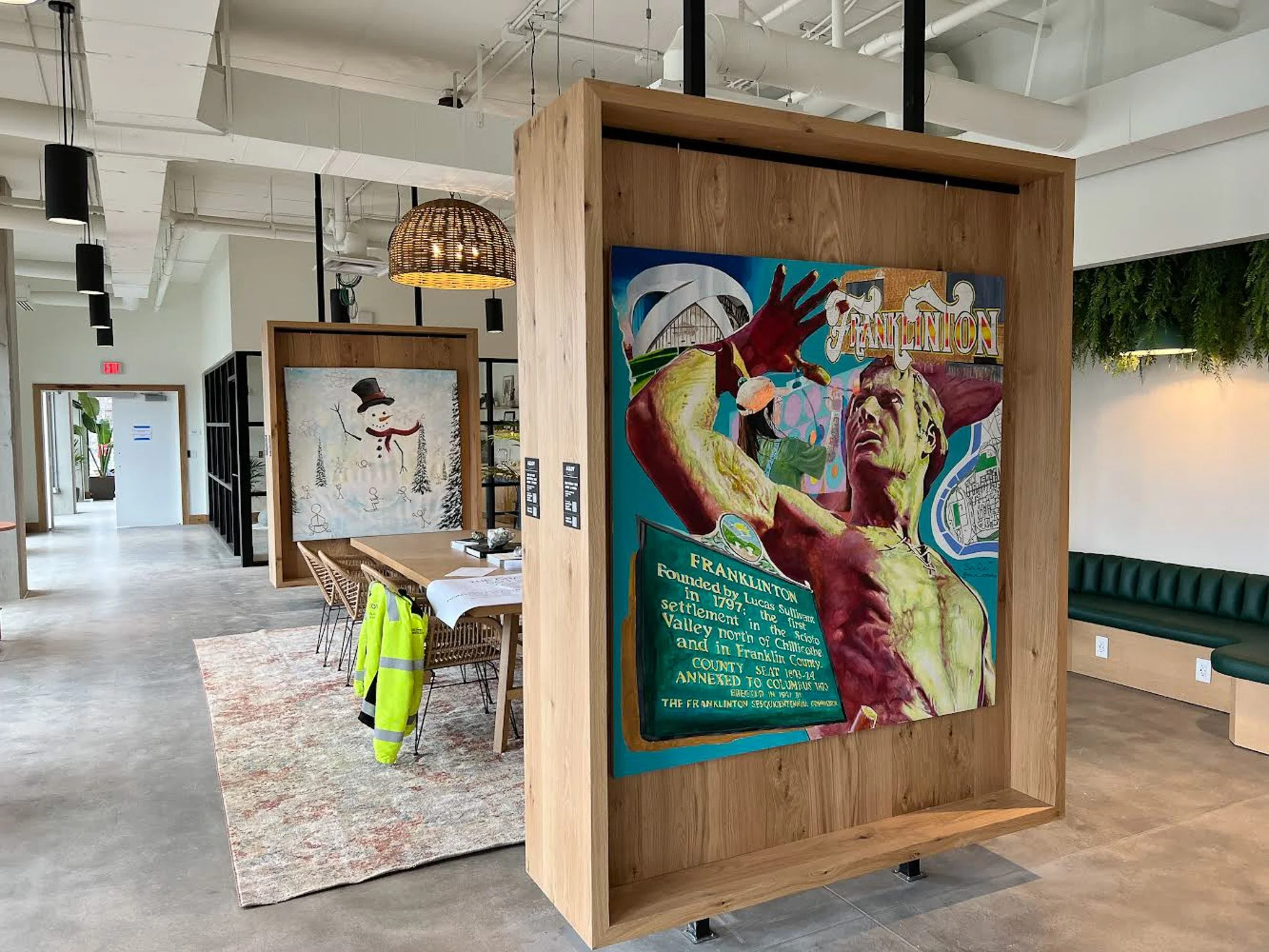 An interior space featuring modern decor with wooden partitions and colorful artwork. In the foreground, a large vibrant painting depicts a figure with exaggerated features and bold colors. Slightly behind, another artwork shows a snowman with a top hat in a winter scene. There is a long dining table with a white tablecloth and chairs, and a high-visibility safety vest hanging over one chair. The space has a polished concrete floor, hanging pendant lights, and a green seating area along the wall.