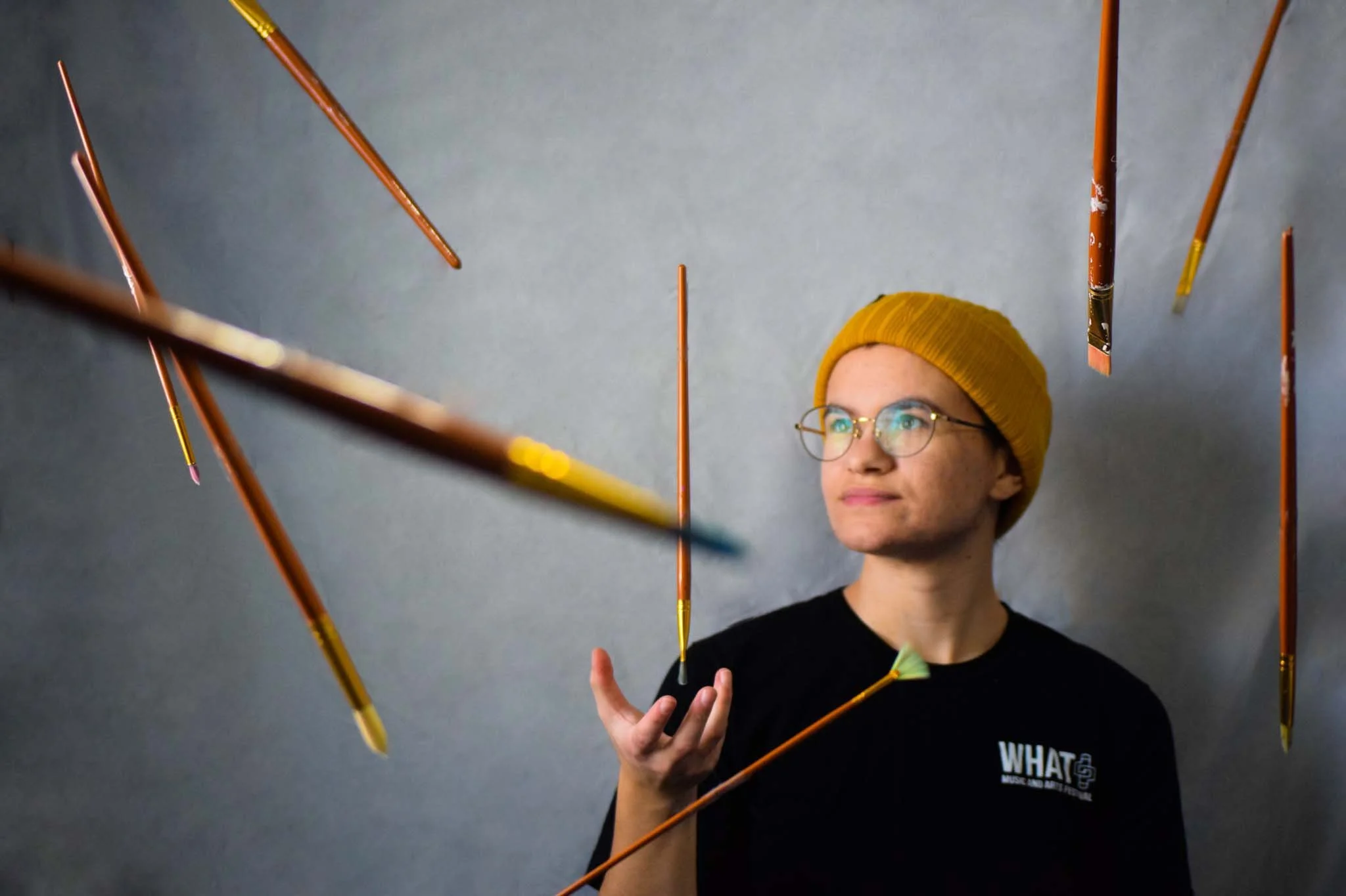 A person with glasses and a yellow beanie, wearing a black shirt, surrounded by floating paintbrushes.
