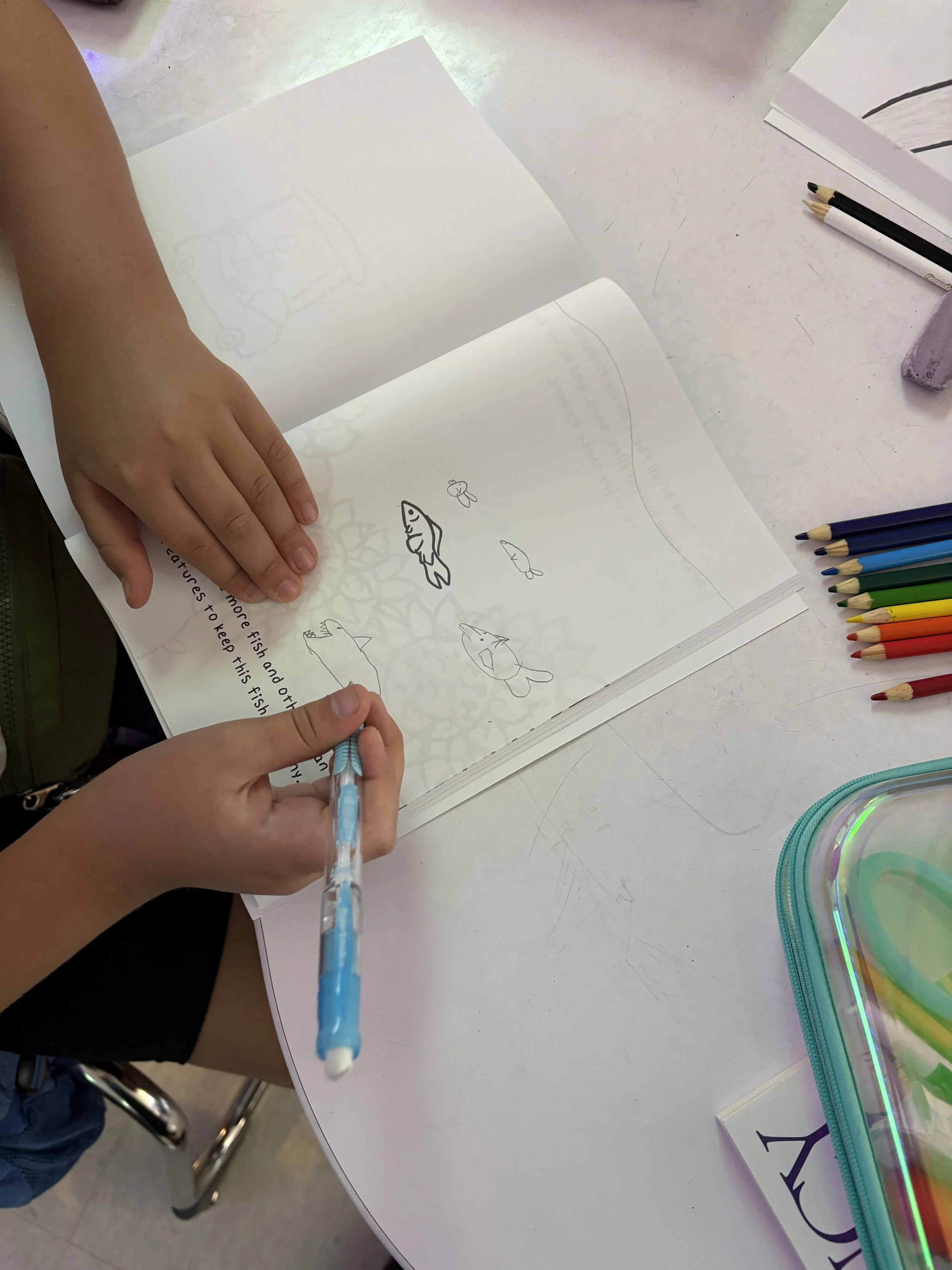 A child drawing fish and other sea creatures in a sketchbook with colored pencils on a white table, with additional art supplies and a purple eraser nearby.