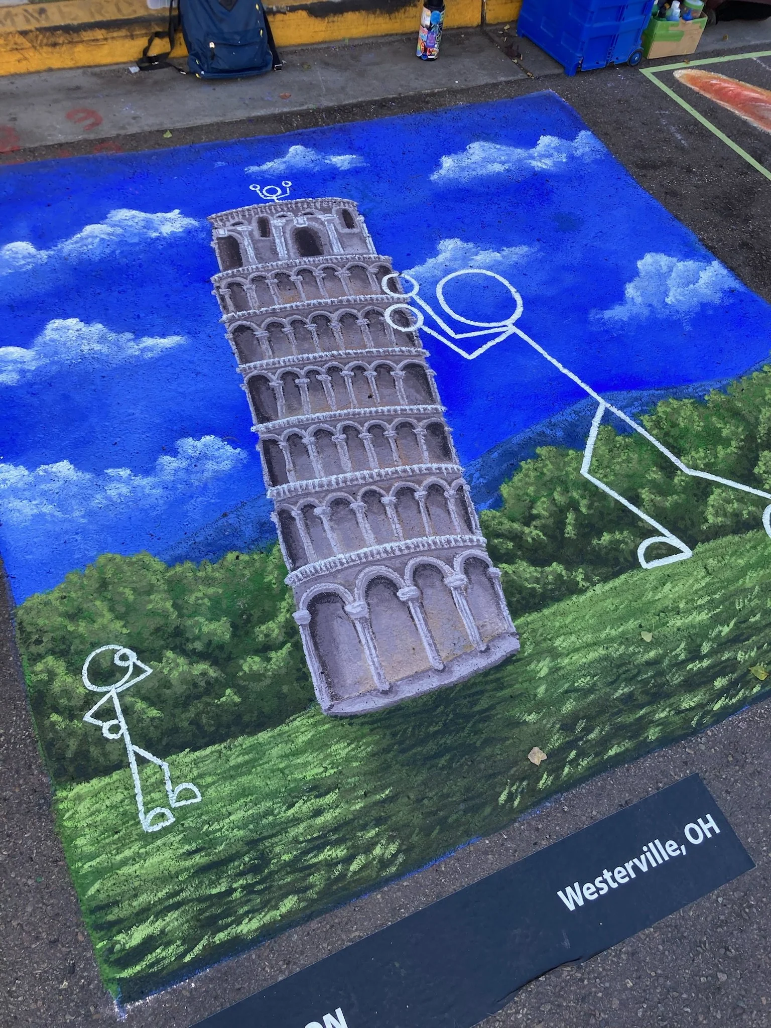 Colorful street painting of the Leaning Tower of Pisa with a stick figure holding up the tower, set against a sky with clouds and green landscape.