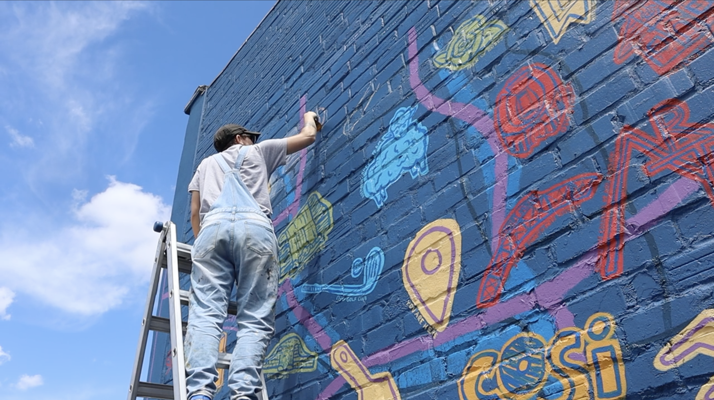 Person on a ladder painting a colorful mural on an exterior brick wall under a blue sky.