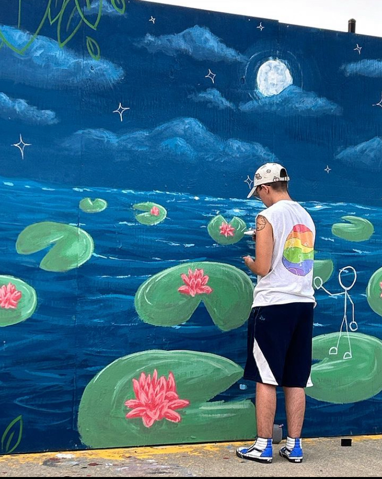 A person standing in front of a colorful outdoor mural featuring a night scene with clouds, a full moon, stars, and a pond with lily pads and pink water lilies.