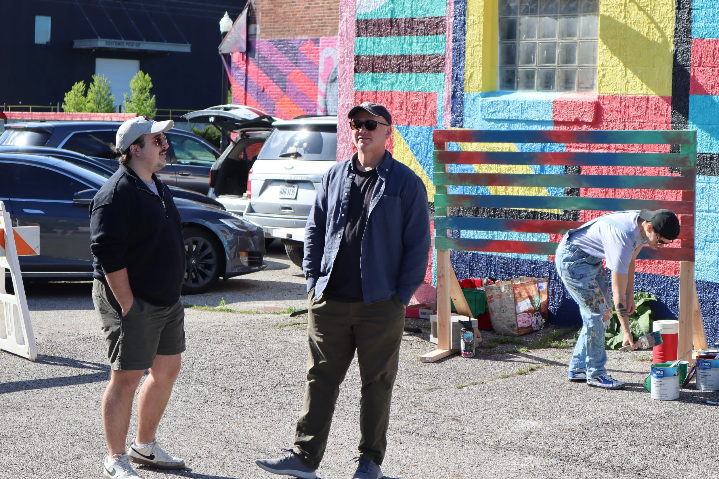 Two men standing and talking in a parking lot with colorful mural walls, while a young man paints a wooden structure.