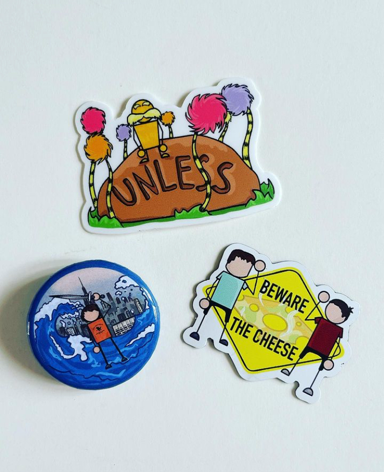 Three stickers on a white surface: one with cartoon creatures in colorful fuzzy hats on a brown rock with the word 'UNLESS'; a round button showing a cartoon person in a boat on water with buildings in the background; and a yellow caution sign with t