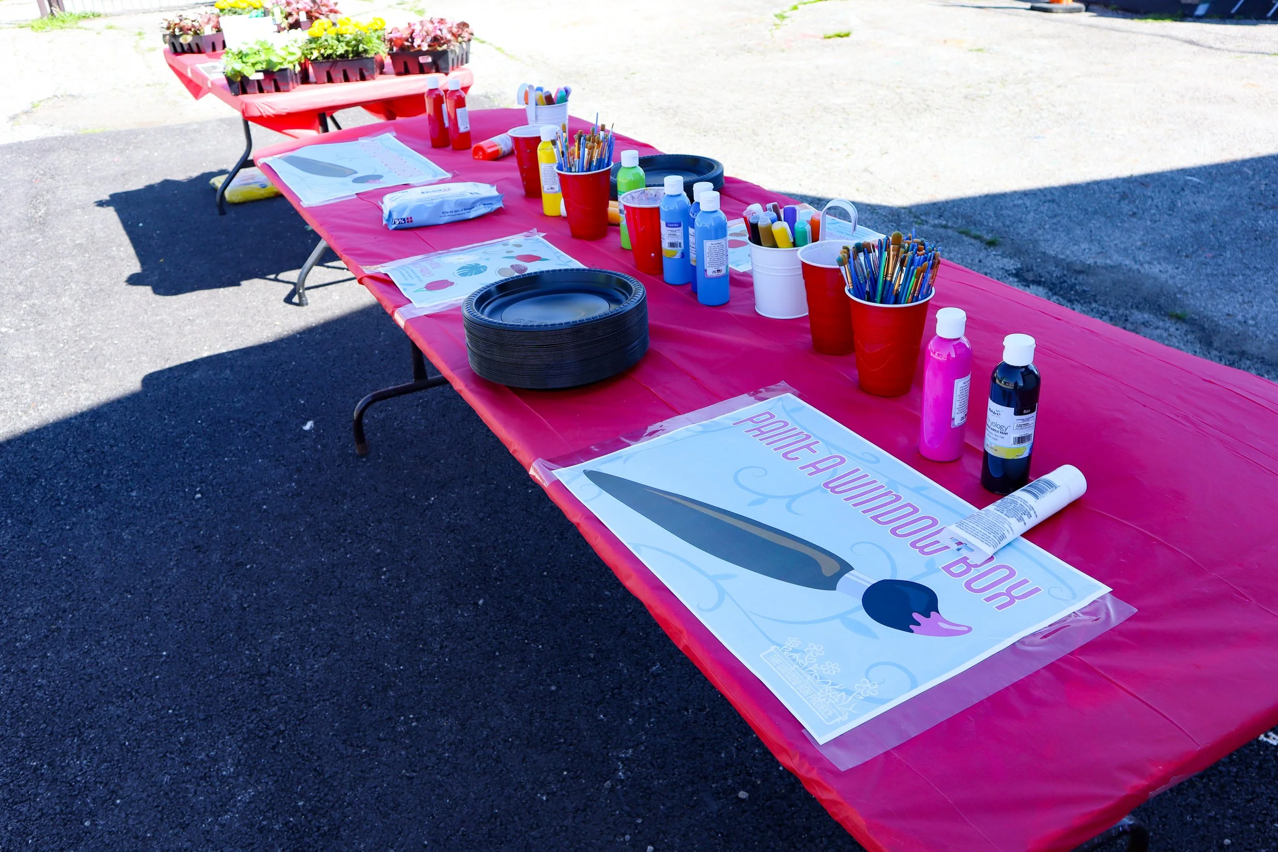 Outdoor table with pink tablecloth set up for a paint-a-witch activity, with painting supplies including paint bottles, brushes, and paper, and a sign that reads 'Paint a Witch' with a witch's hat illustration.