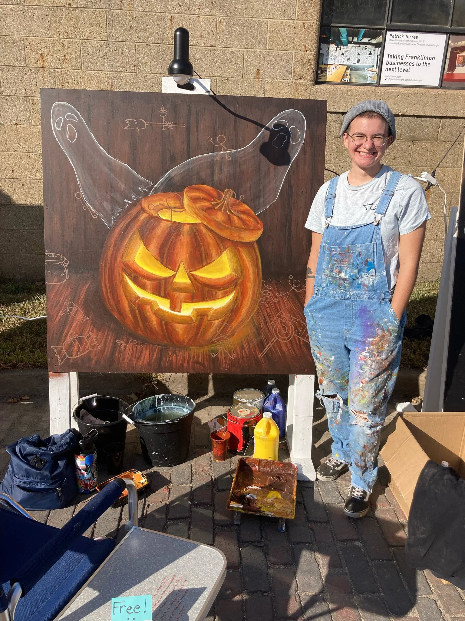 A young artist wearing overalls and a beanie is standing next to a large Halloween-themed pumpkin painting on a canvas. The pumpkin has a carved face with a mischievous smile and glowing eyes. The artist's paint-splattered clothes and the paint supplies on the ground indicate she has been working on the artwork outside.