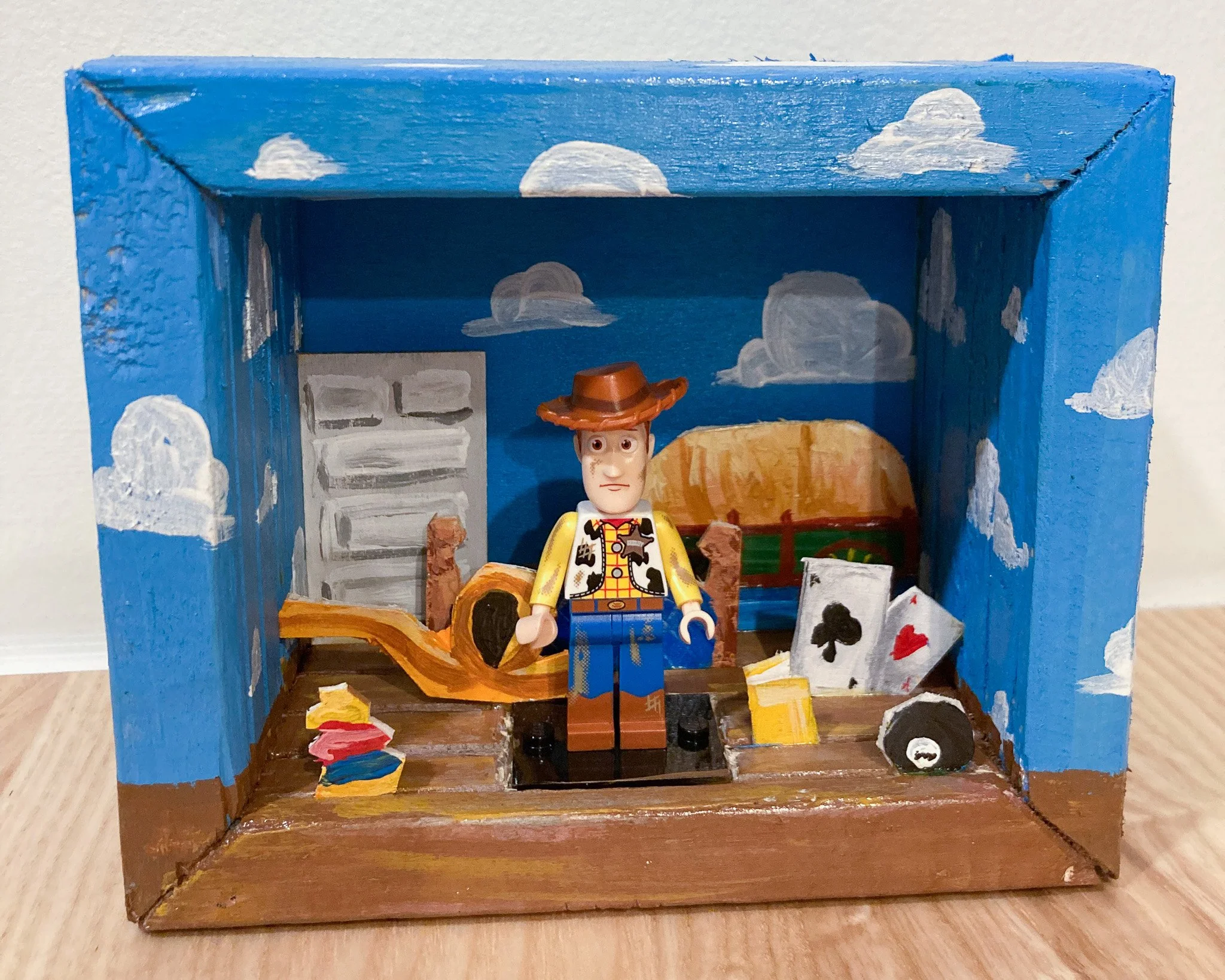 A toy diorama of a cowboy in a wooden box with painted blue sky, white clouds, and a desert background. The scene includes playing cards, a guitar, a small speaker, and other miniature items.