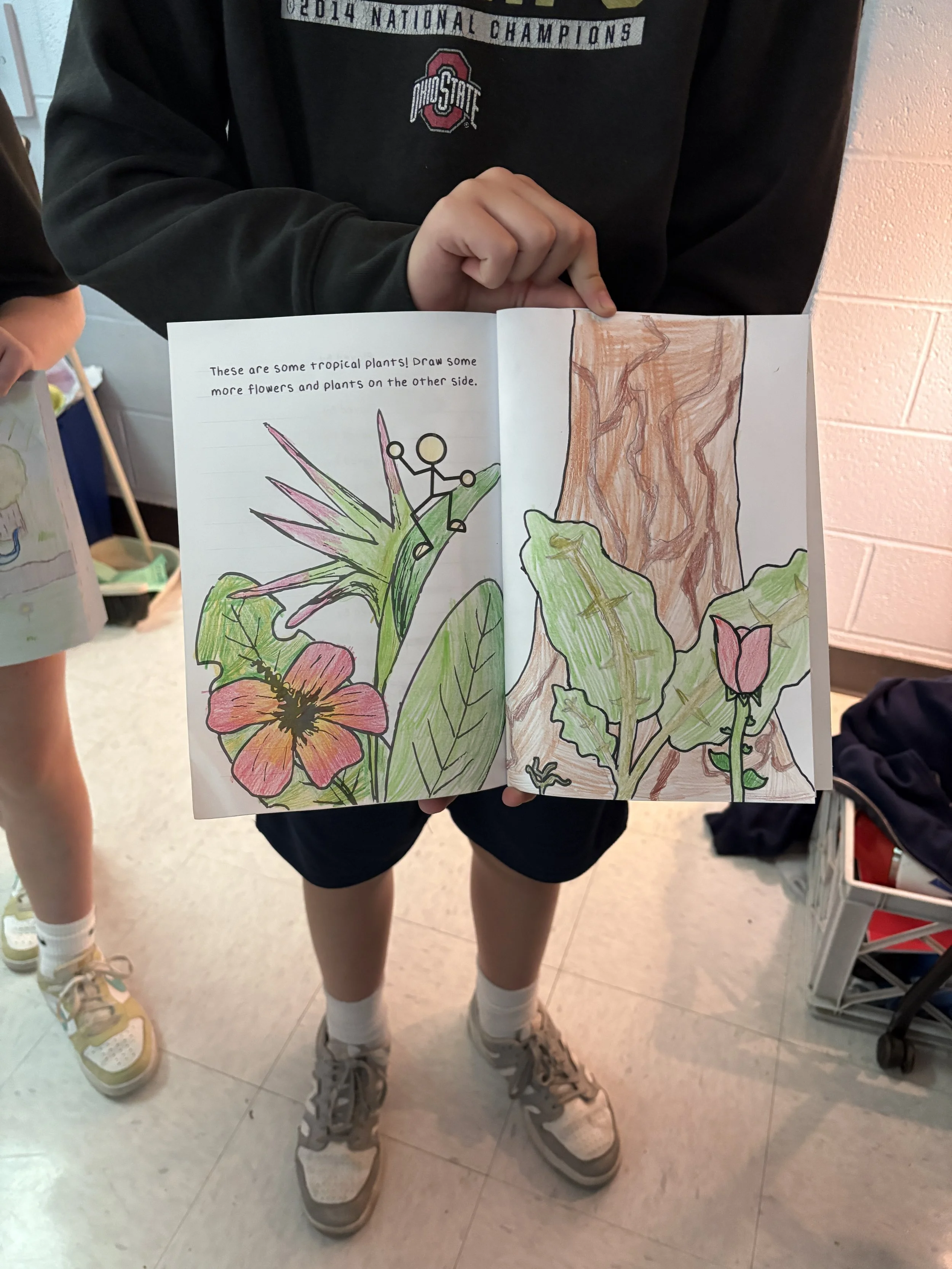 Child holding open coloring book with tropical plants and flowers, including a pink flower and green leaves, and a drawing of a brown tree trunk on the right side.