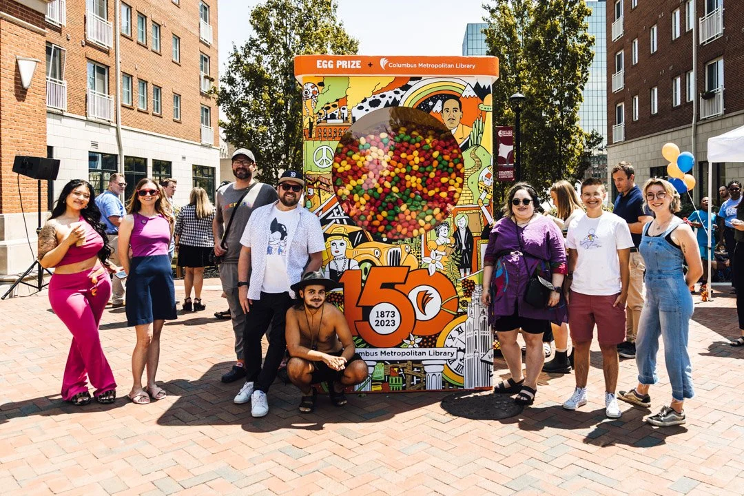 Group of people celebrating outdoors near a large vending machine holding easter eggs, commemorating 150 years of the Columbus Metropolitan Library.