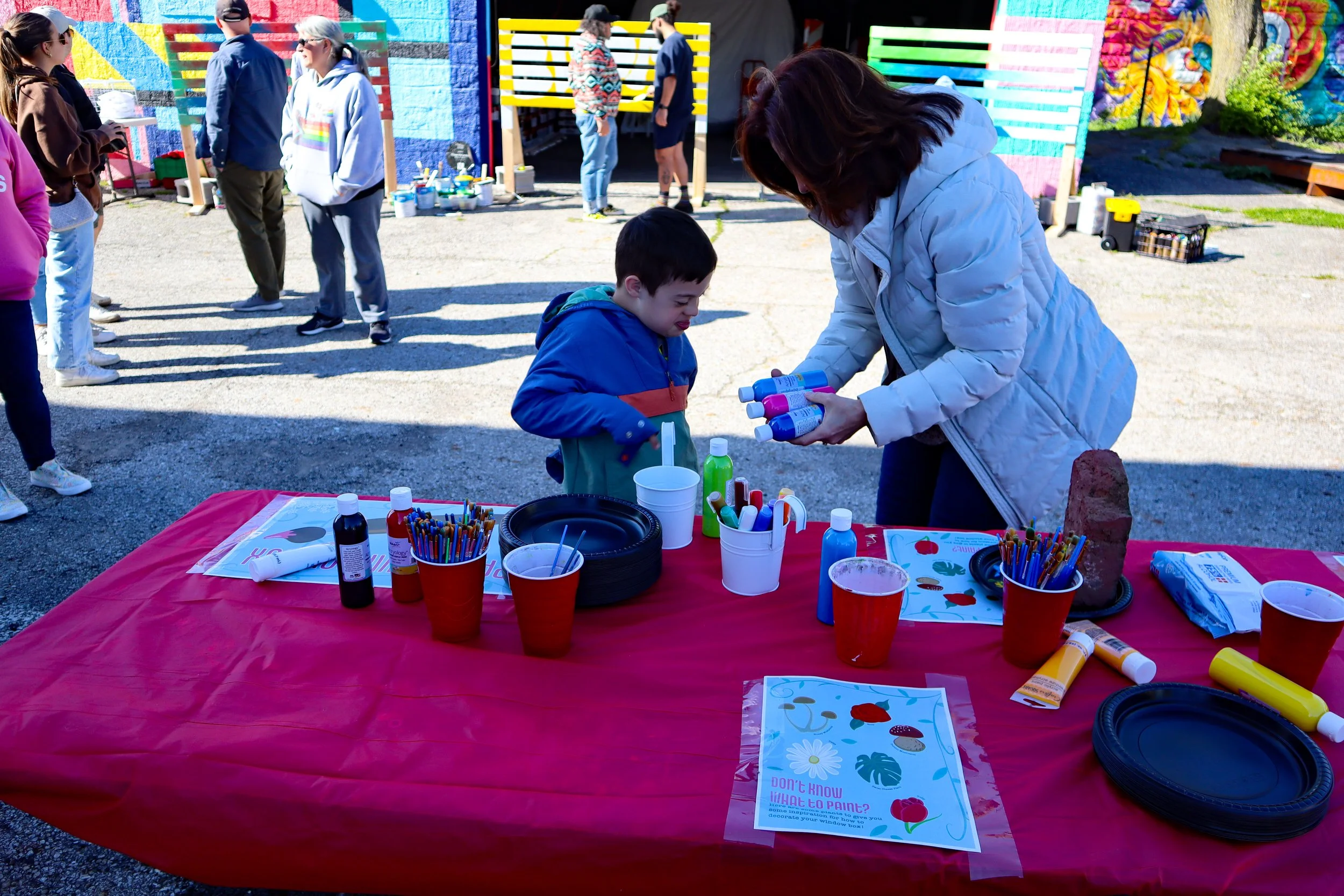 A woman and a young boy at a colorful outdoor art event. The woman is handing bottles of paint to the boy, who is looking at them. The table in front of them has art supplies like paint bottles, brushes, and paper. In the background, several people are gathered, and there is a mural painted on the wall.