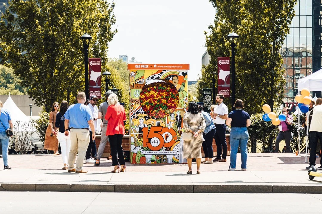 People gathered around a colorful vending machine commemorating the 150th anniversary of the Columbus Metropolitan Library, with trees and tents in the background.