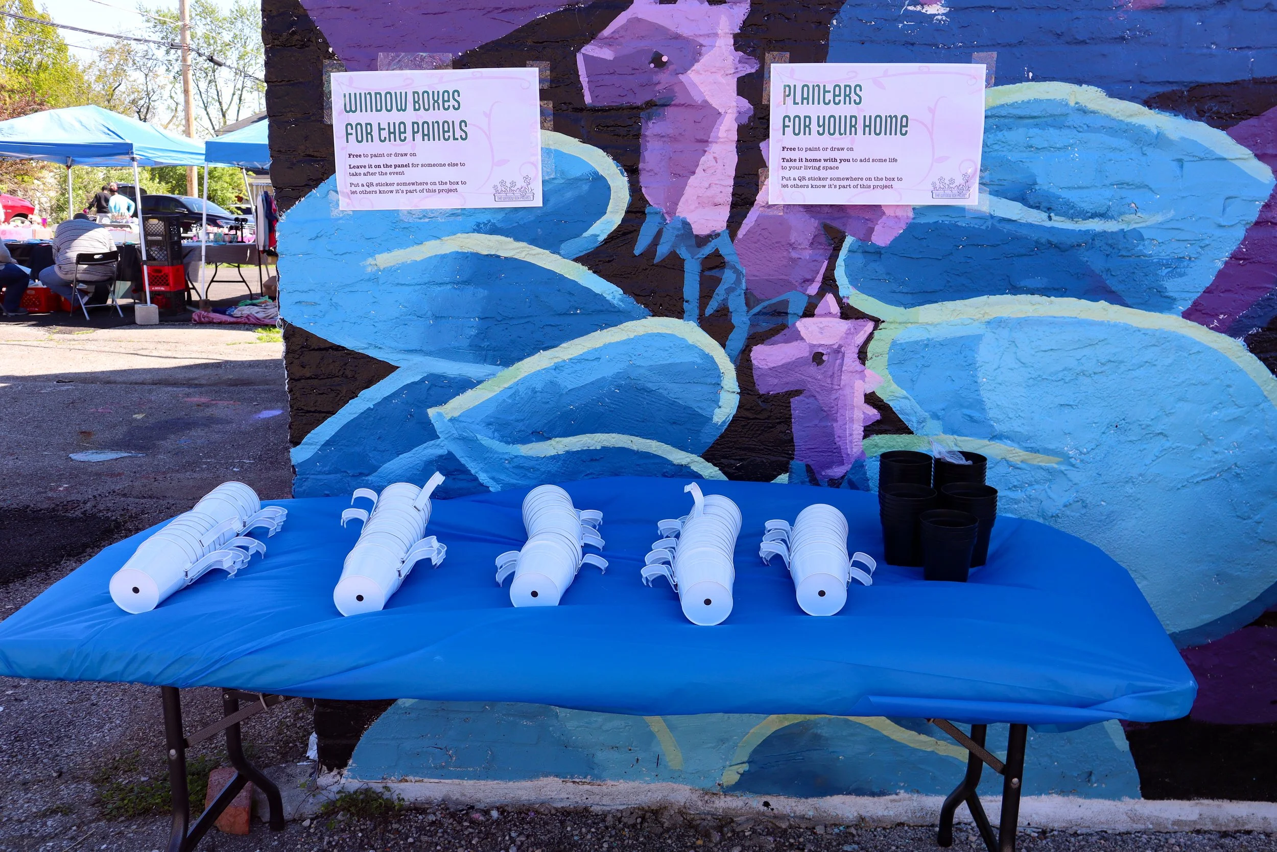 Table with white and black plastic containers shaped like lobsters, set in front of a colorful mural on a brick wall. The mural features abstract blue and purple shapes, possibly representing a woman's face with flowing hair. Two signs are posted on the wall, one about window boxes for panels and another about planters for your home.