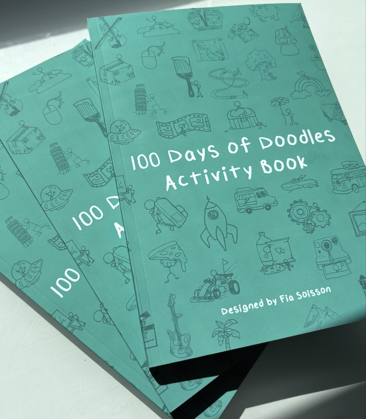 Three teal-colored activity books titled "100 Days of Doodles Activity Book" designed by Fia Soisson, featuring doodle illustrations of various objects like a rocket, guitar, pizza, and trees.
