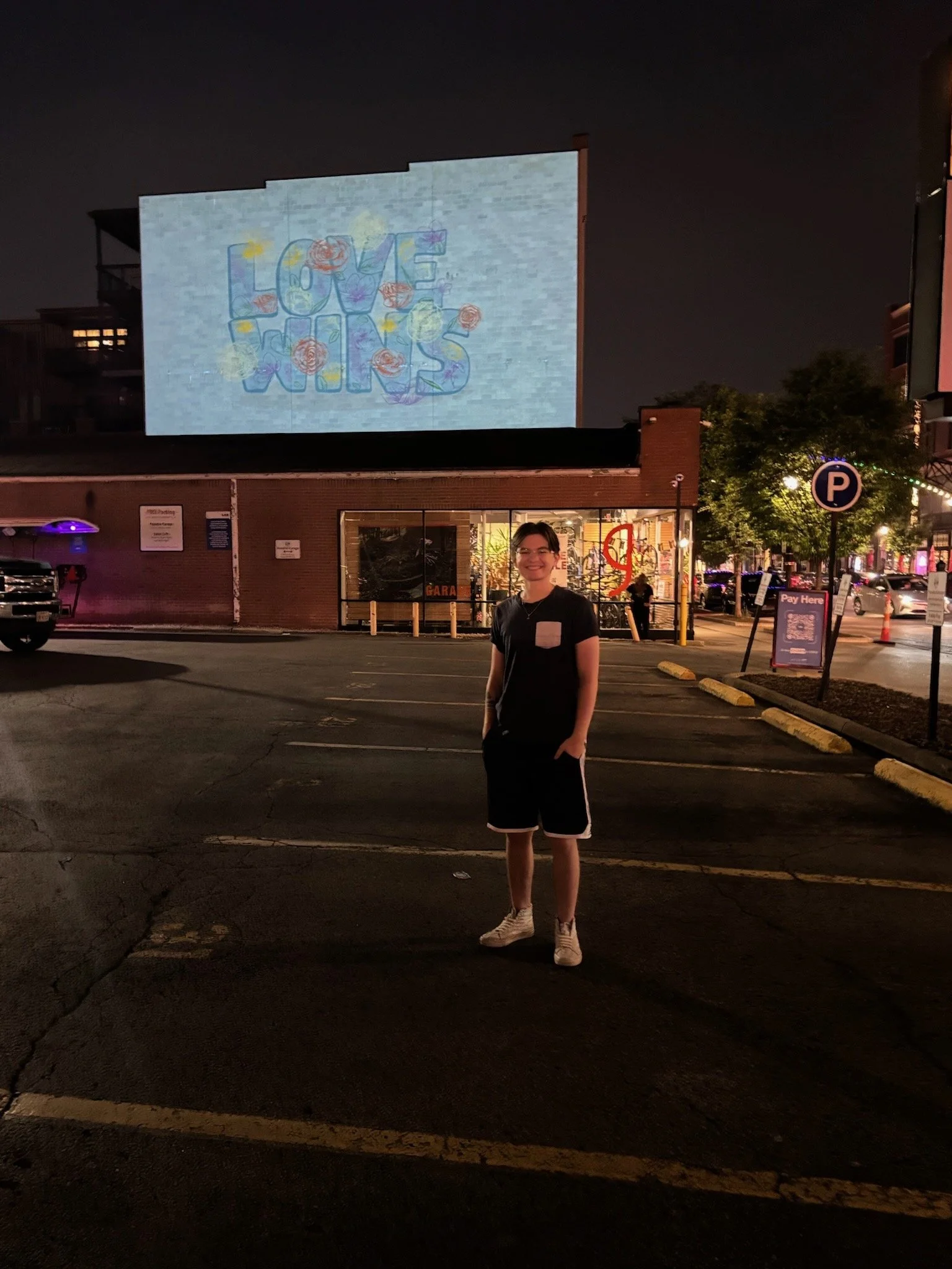 A person stands in a parking lot at night in front of a brick building with a large illuminated screen projecting the words 'LOVE WINS' decorated with flowers.