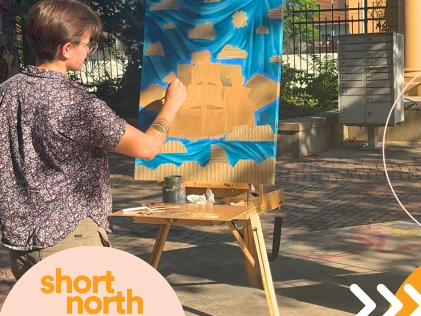 An artist painting a mural on a canvas outdoors. The mural depicts a stylized mountain landscape with a blue sky and clouds. The artist is standing to the left, wearing glasses and a patterned shirt, working with a brush. A small table holds paint supplies including jars and brushes, and the area is surrounded by a metal fence, with trees and a brick path visible in the background.