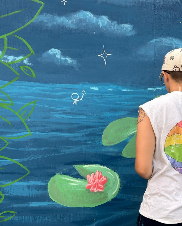 Person standing in front of a colorful mural of a pond with lily pads, a pink water lily, and a sky with clouds and stars.