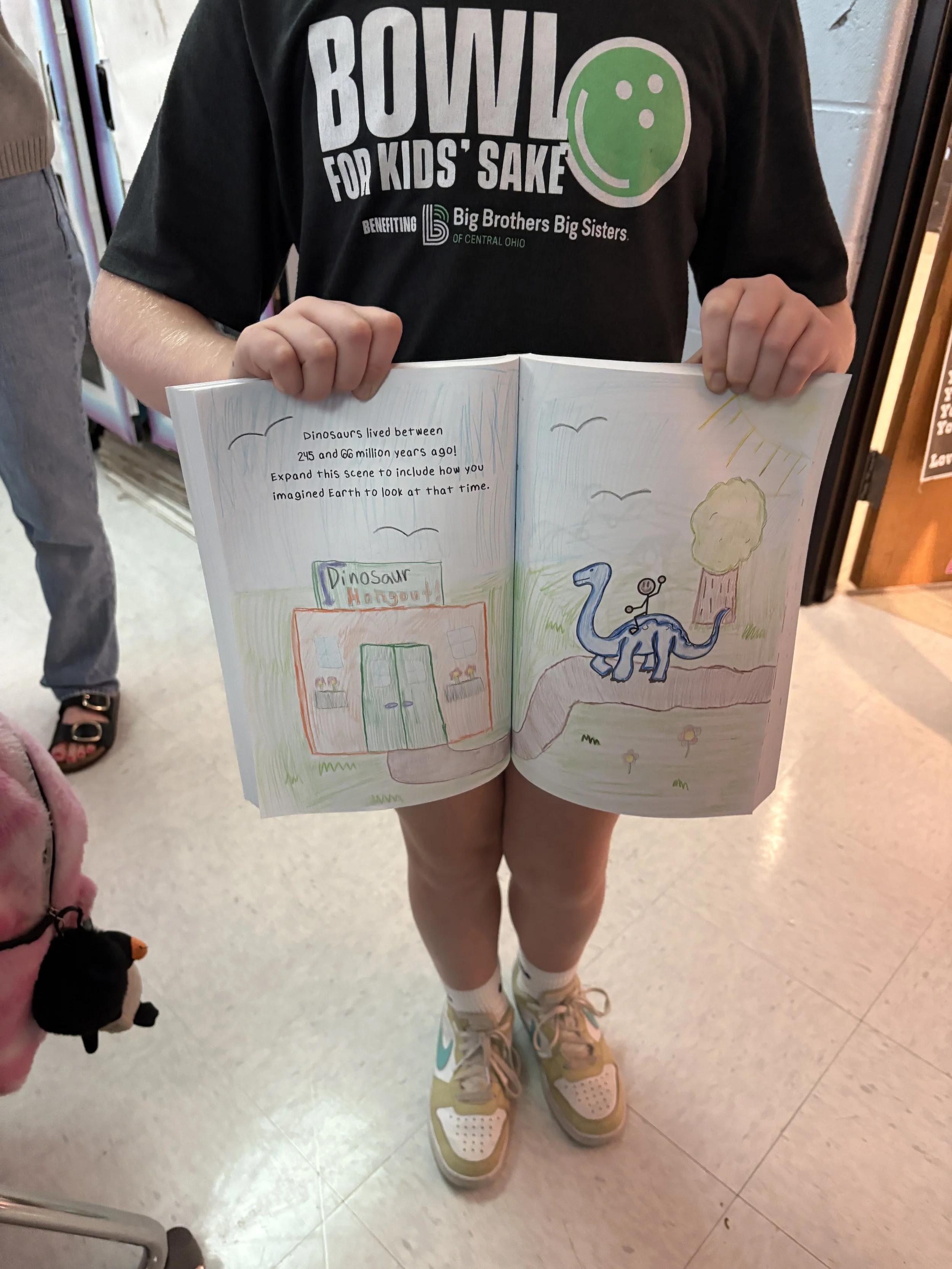 A child wearing a black t-shirt with a smiley face graphic, holding an open drawing book. The drawing depicts a scene of dinosaurs, a dinosaur hangout building, and a sunny outdoor landscape with a tree and grass.