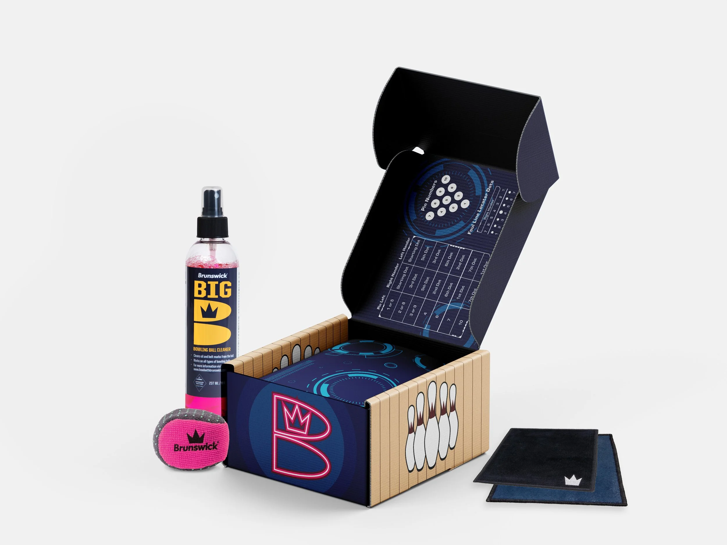 A bowling ball cleaning kit includes a spray bottle labeled 'Brunswick Big Bowling Ball Cleaner,' a pink and gray Brunswick cleaning sponge, a black textured cloth, and an open box with bowling pin images and game instructions inside.