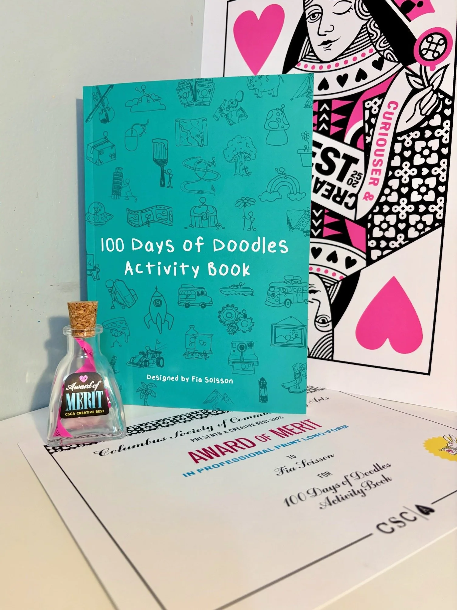 A certificate and a small glass bottle with a cork contain award titles, displayed next to a blue activity book titled "100 Days of Doodles Activity Book" on a white surface. The certificate is from Columbus Society of Creatives and is awarded to Fia Soisson for the activity book.