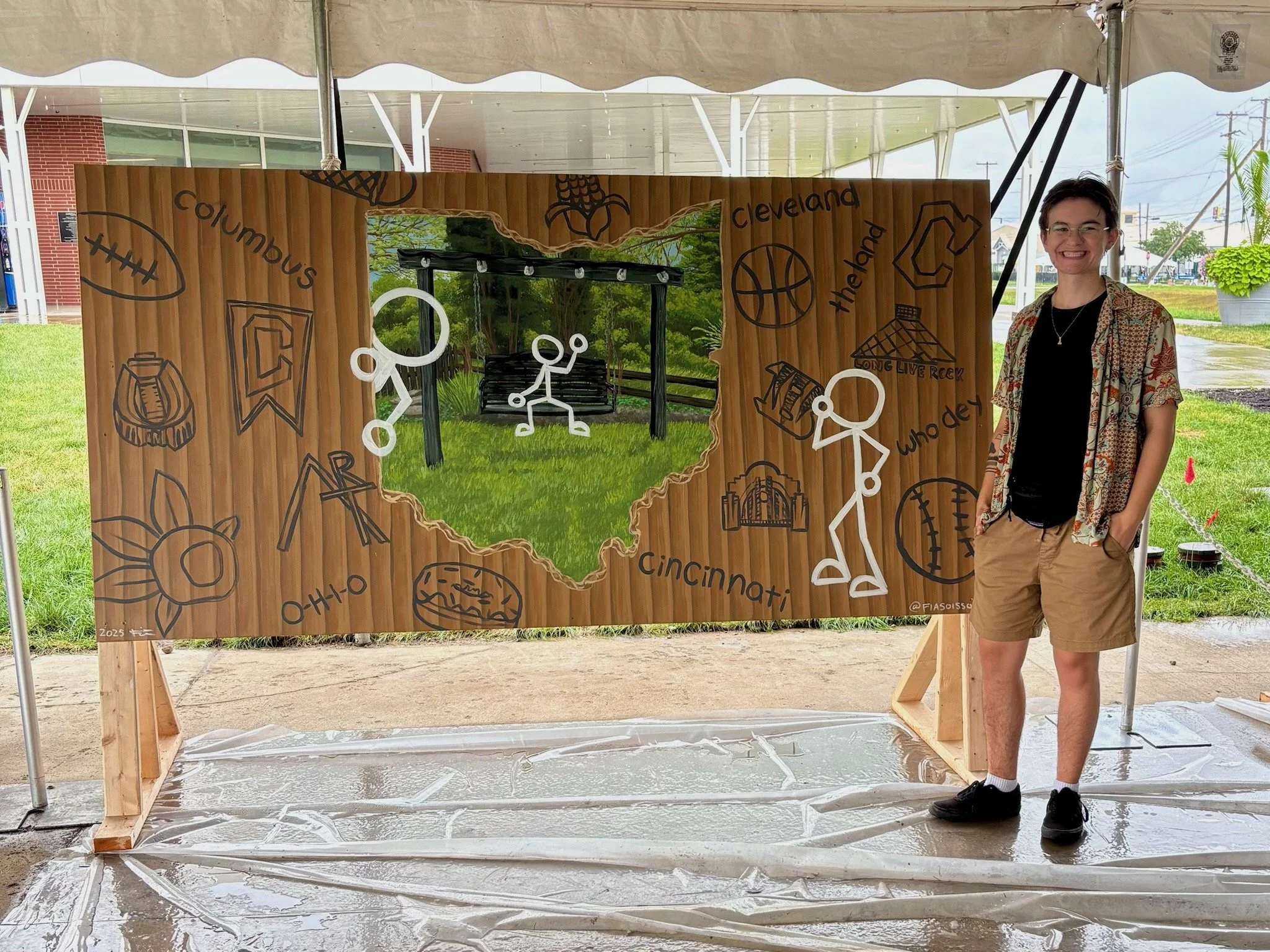 A person standing next to a large wooden board decorated with drawings of Ohio cities, sports symbols, and other icons, with a cut-out in the shape of the state of Ohio showing a park scene with a swing and greenery.