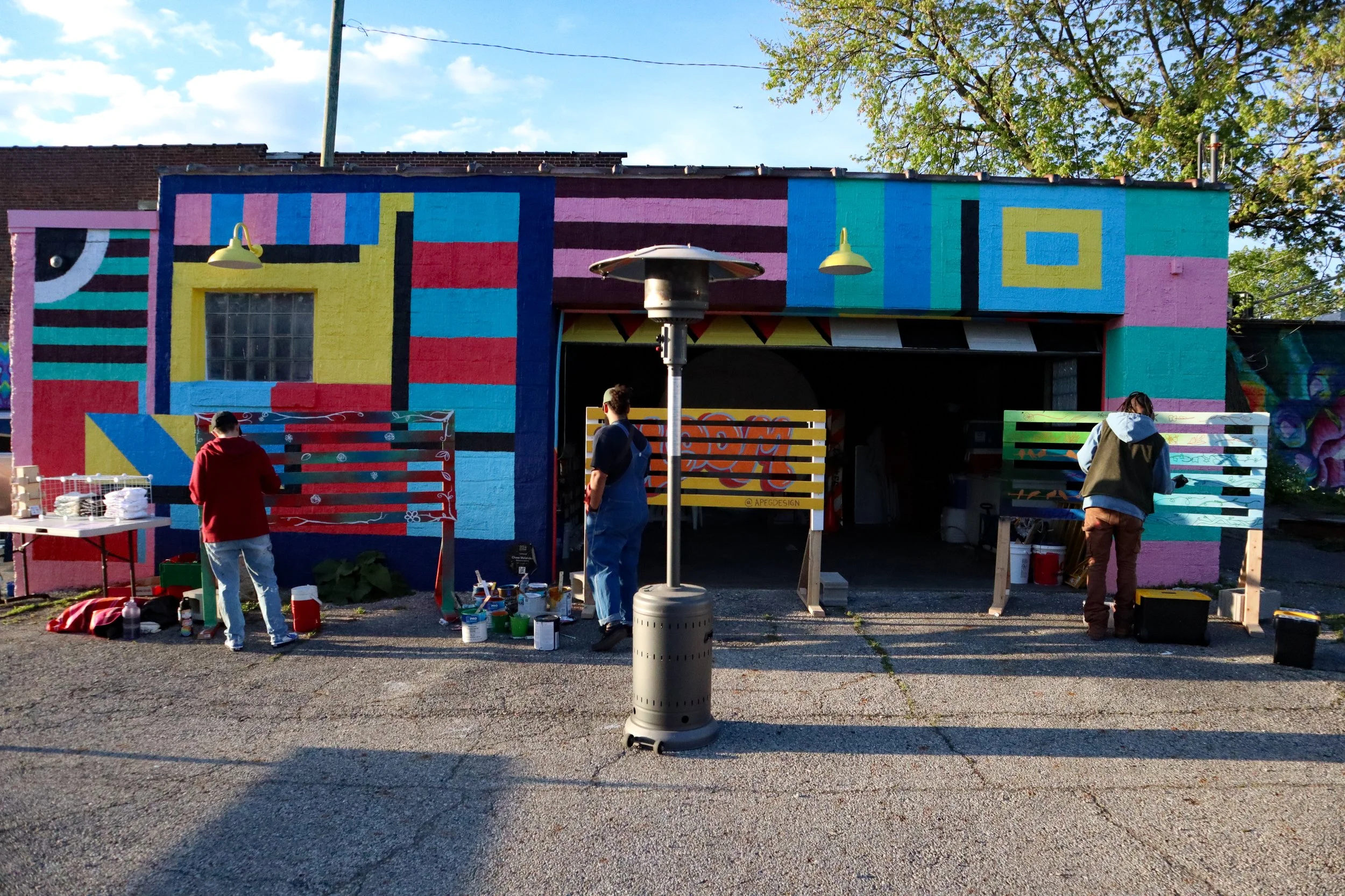 Three people painting a colorful mural on a building with a pink, yellow, red, blue, and green pattern, set against a partly cloudy sky and trees.