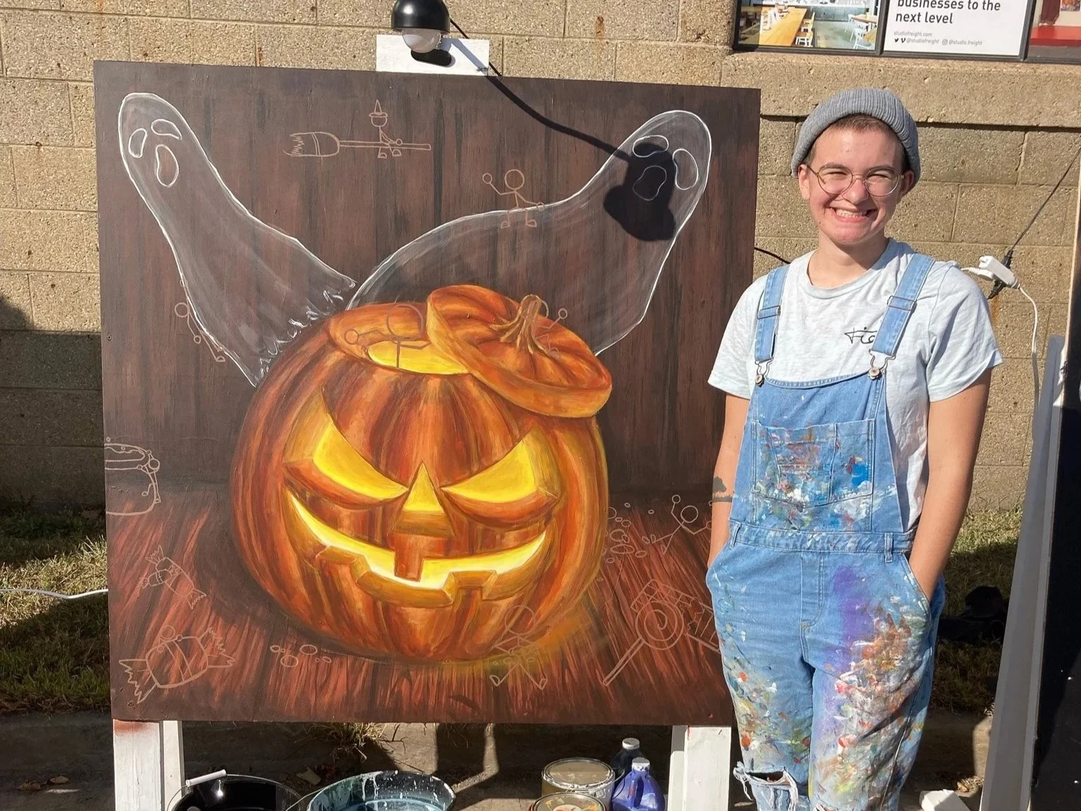 An artist standing next to a chalk art painting of a carved Halloween pumpkin with a mischievous face. The pumpkin is painted in vibrant orange and yellow tones, and a ghostly figure with outstretched arms appears to emerge from the top. The artist is smiling, wearing a gray beanie, glasses, a gray t-shirt, and blue overalls splattered with paint.