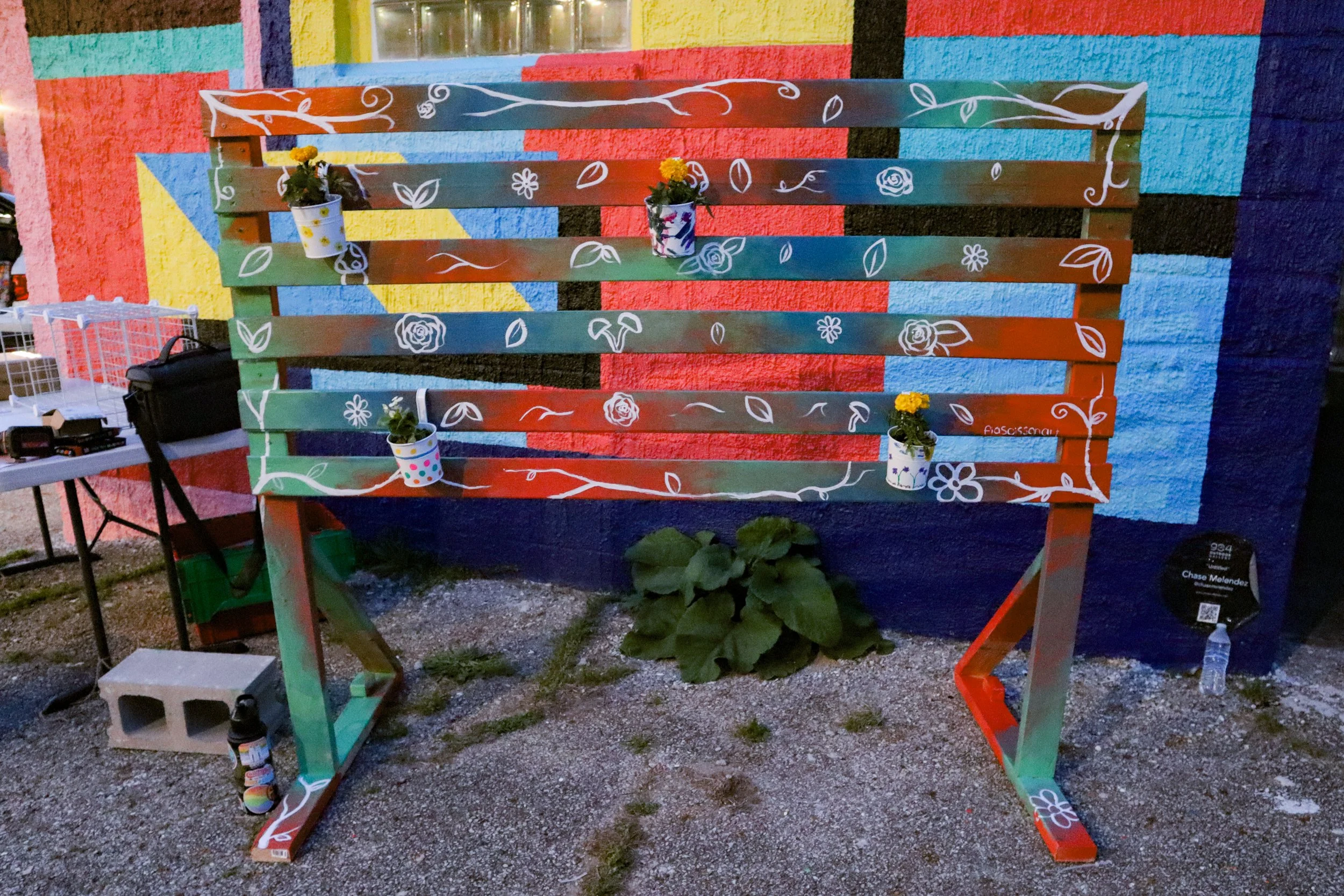 Colorful wooden bench with white floral designs painted on it, placed against a vibrant multicolored mural wall. Four small pots with yellow flowers are attached to the bench.