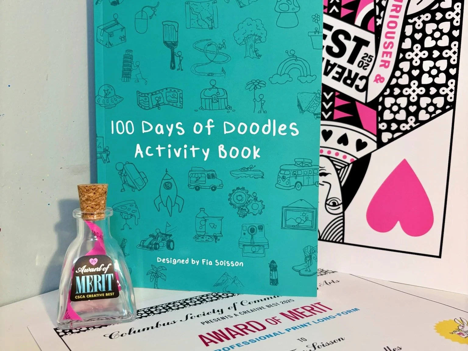 A small glass bottle with a cork stopper, labeled 'Award of Merit' and 'CSCA Creative Best,' placed on a white surface. Behind it is a turquoise activity book titled '100 Days of Doodles Activity Book' designed by Fia Soisson, with illustrations of various doodles. To the right is a card with bold black, white, and pink patterns and the words 'Award of Merit' and 'Professional Print Long-Form' partially visible.