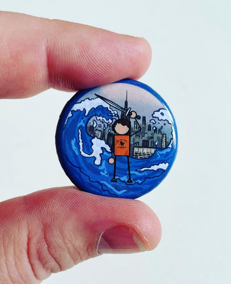 A round pin with a cartoon illustration of a person standing on water with large waves, holding a sword, with a city skyline and mountains in the background.