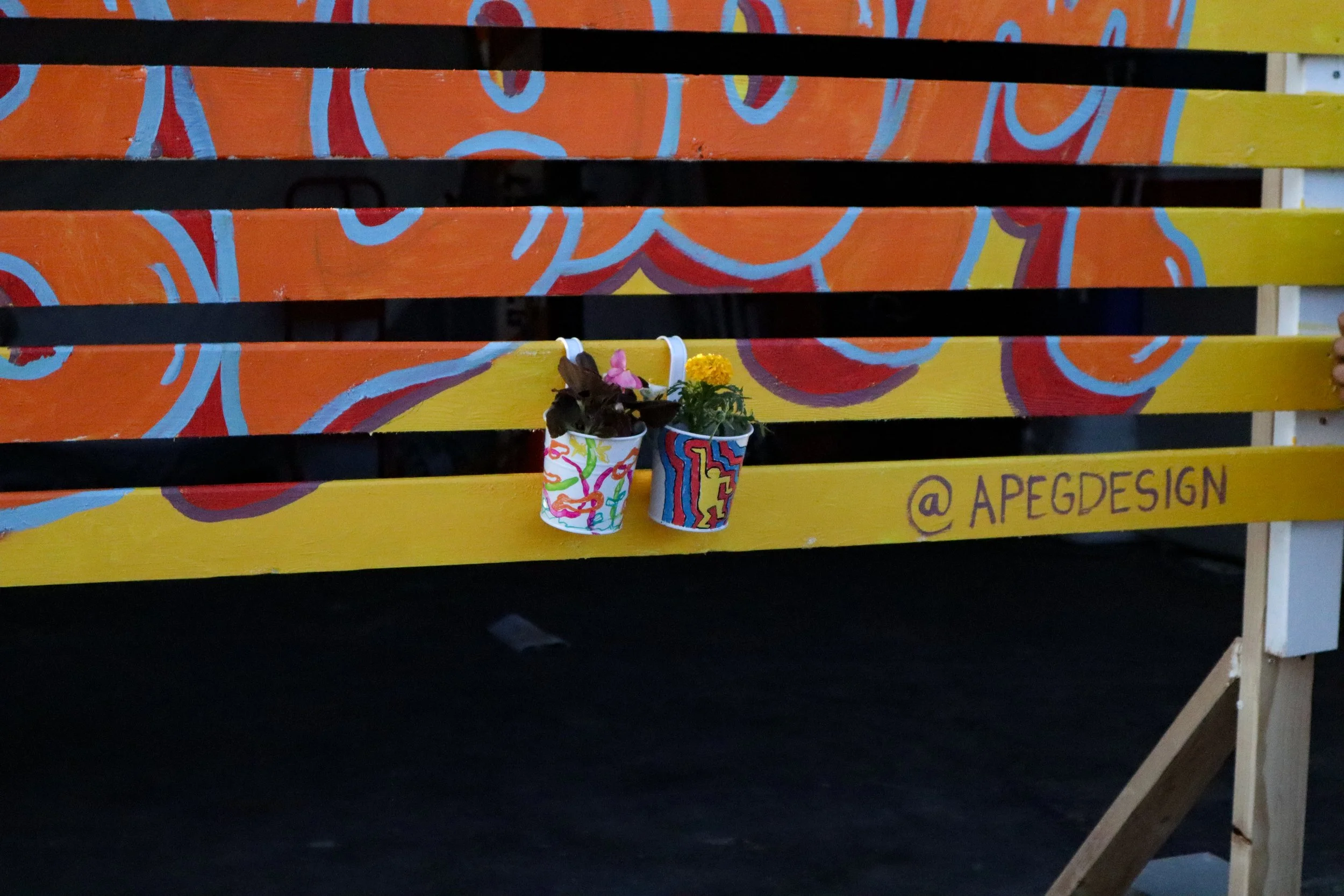A colorful wooden bench painted with abstract patterns in orange, red, yellow, and blue. Two small decorated pots with flowers and plants are placed on the bench, with one pot having a butterfly decoration. The bench has the Instagram handle '@APEGDESIGN' written in purple on one of the yellow slats.