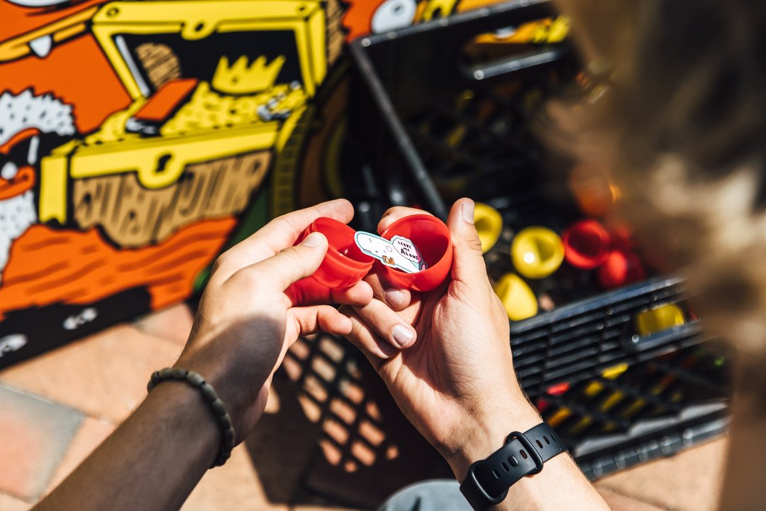 A person holding a red container with a sticker that says 'Build Your Own' near a basket of yellow and red bottles or containers, with a colorful cartoon-style background.