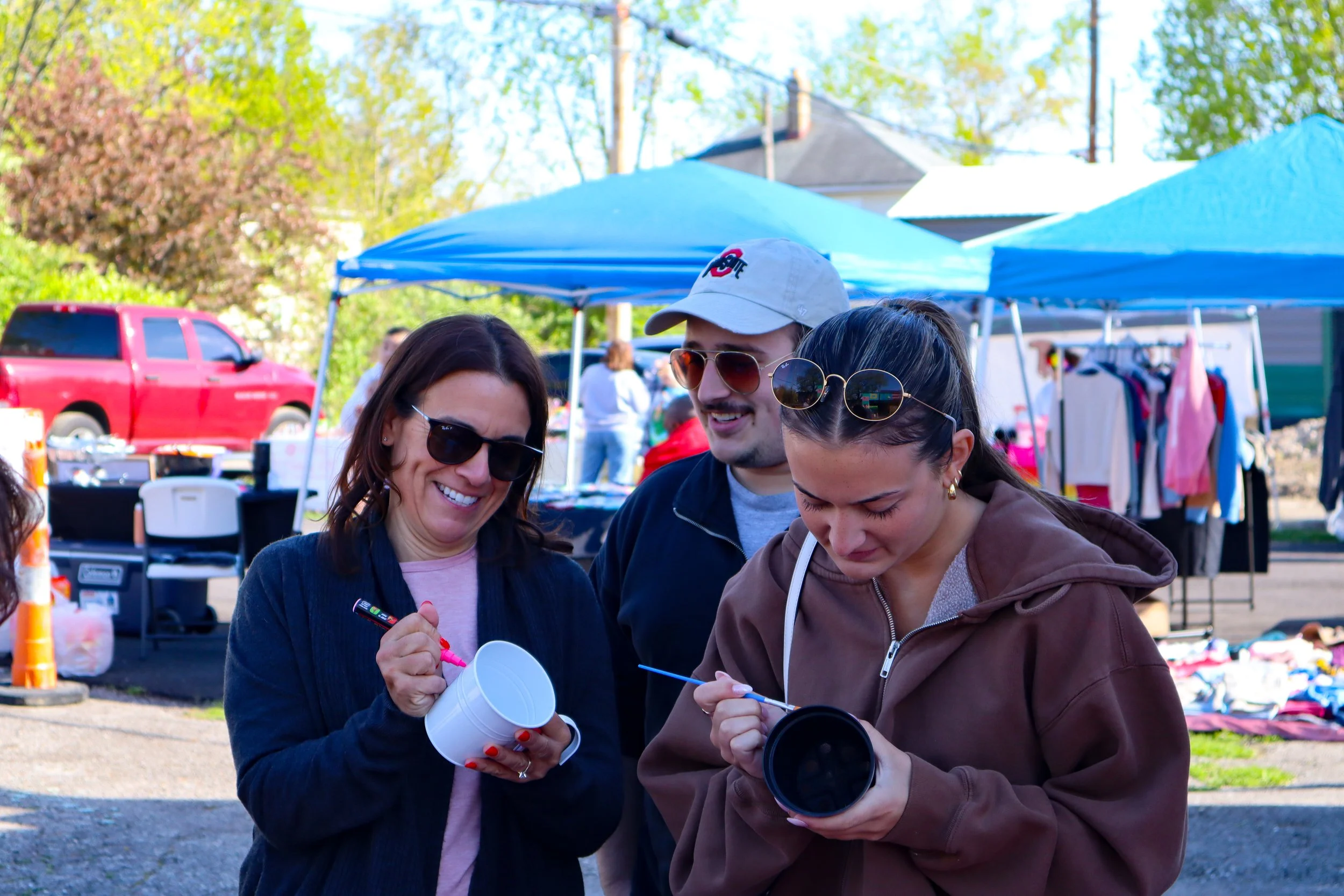 Three people are smiling and looking at a camera lens, at an outdoor yard sale with blue tents, clothing racks, and a red truck in the background.