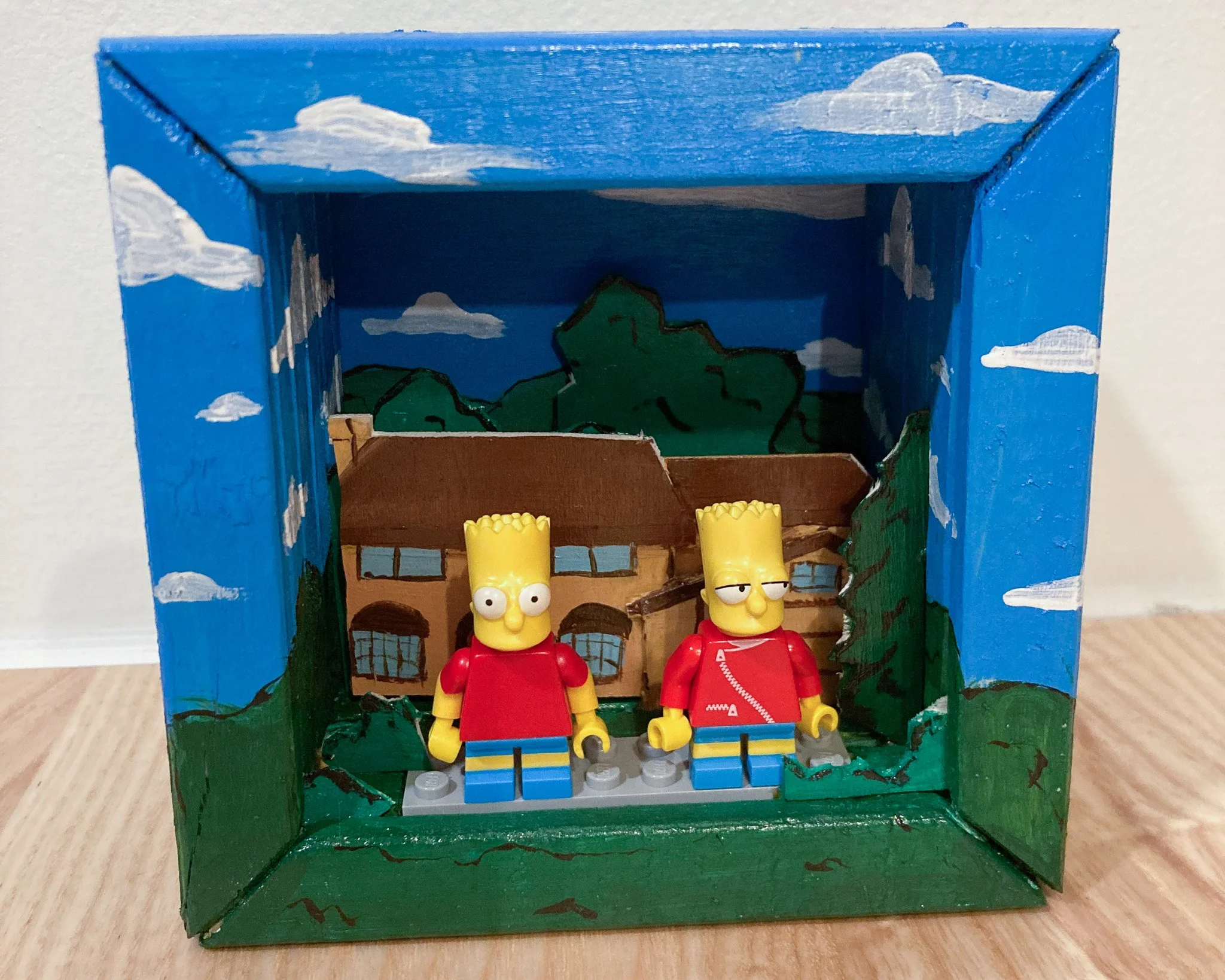 A shadow box craft depicting a scene from 'The Simpsons' with two LEGO figures of Bart Simpson and another character, standing in front of a painted background of trees and a house.