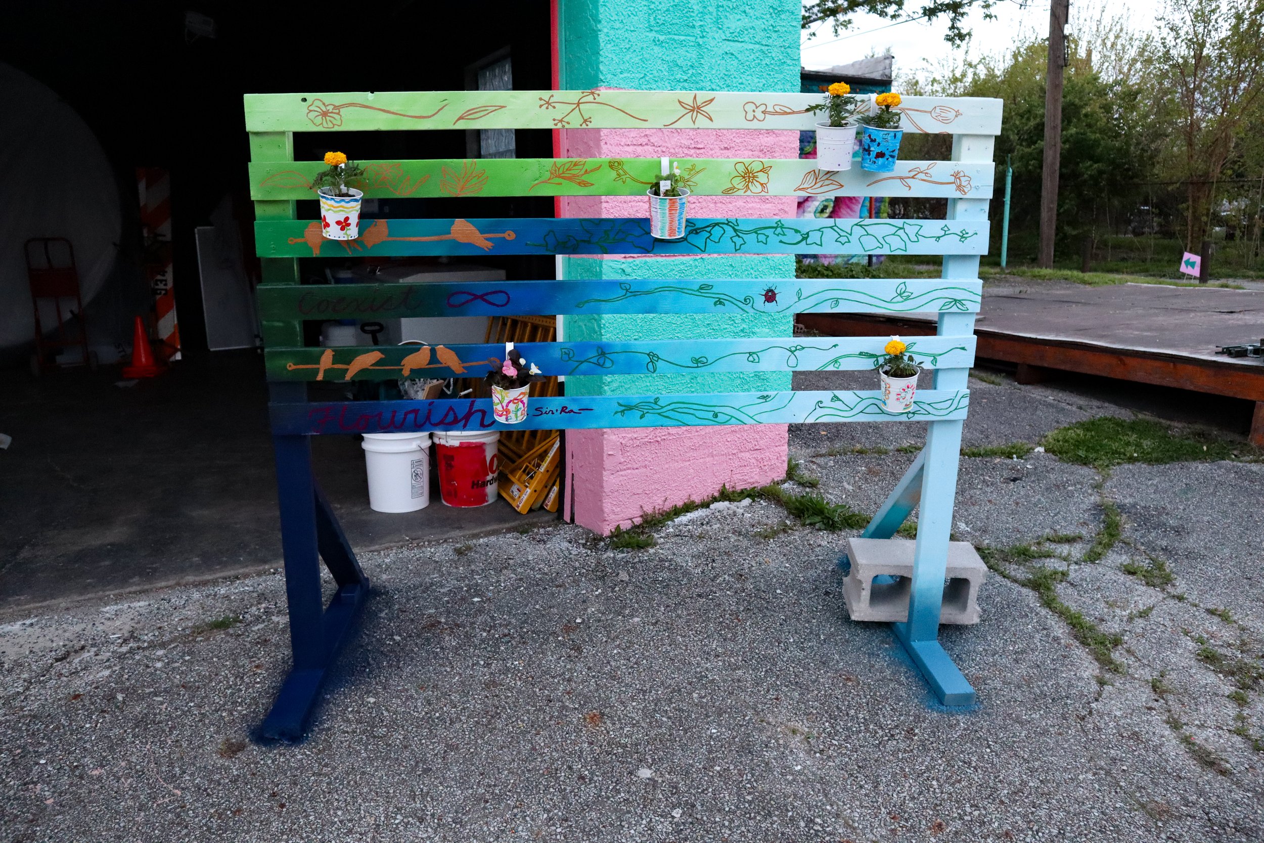 Colorfully painted wooden panel with small flower pots with orange, pink, and yellow flowers attached to it, located outdoors on dirt ground.