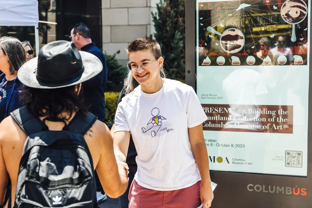 A person with glasses and a white T-shirt shaking hands with a person wearing a black hat and backpack outdoors, with an informational poster for an art exhibit in the background.