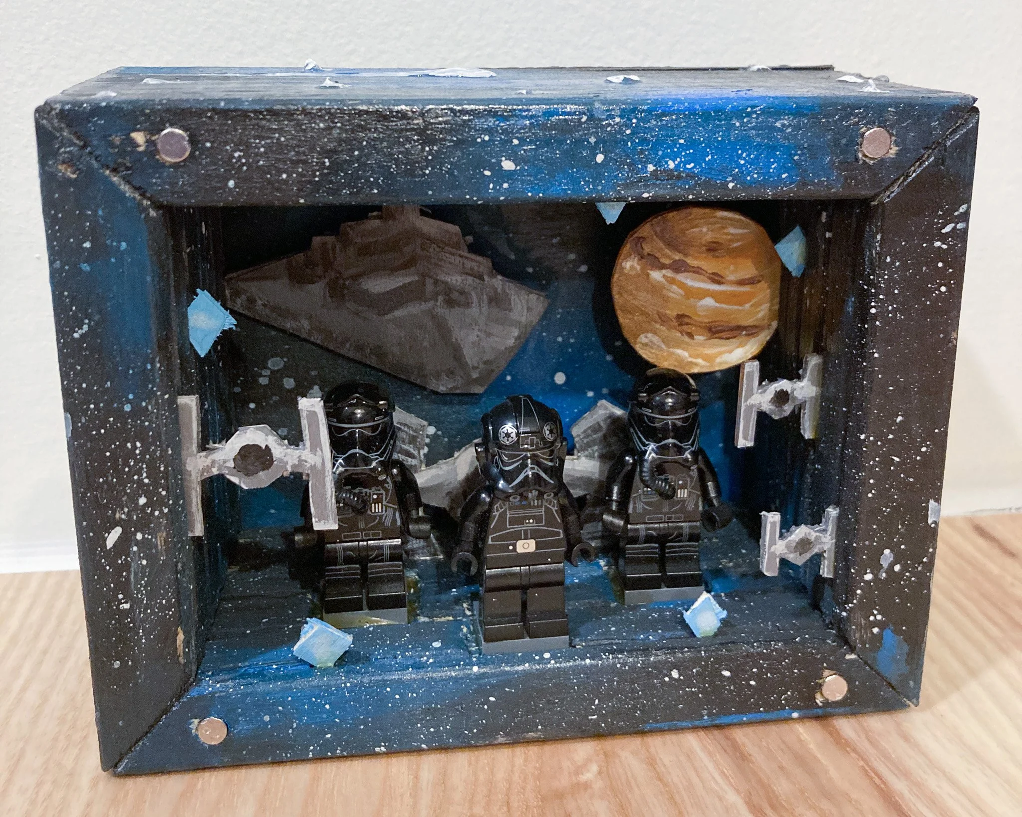 Star Wars themed display with three Lego stormtroopers inside a black box decorated with space and planetary background, including a planet and a moon, with painted splatters and a small model of the Death Star.