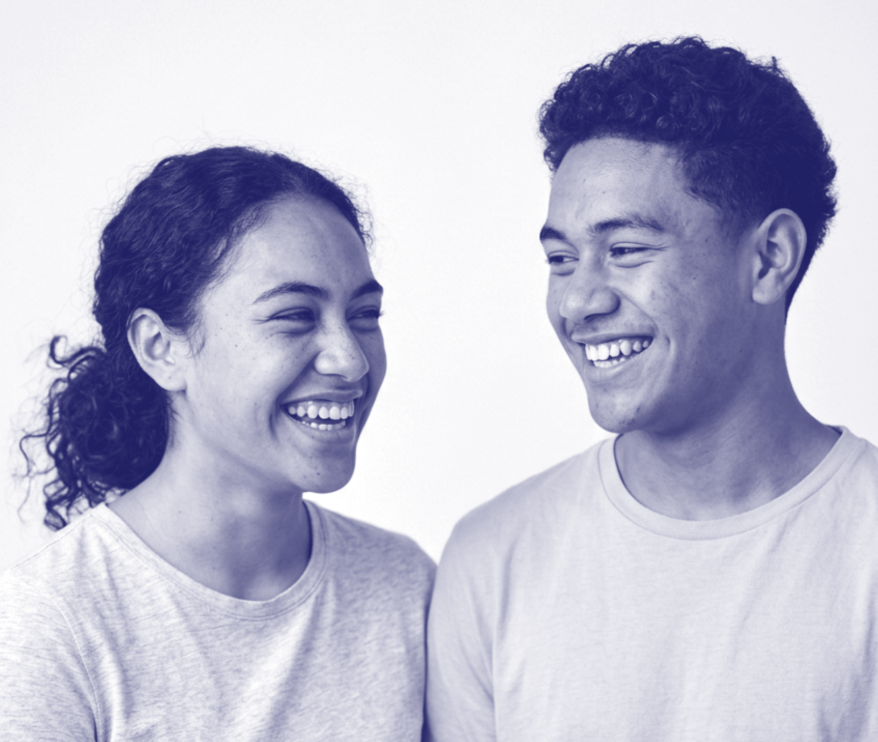 Two young adults, a woman and a man, smiling and looking at each other against a plain background.