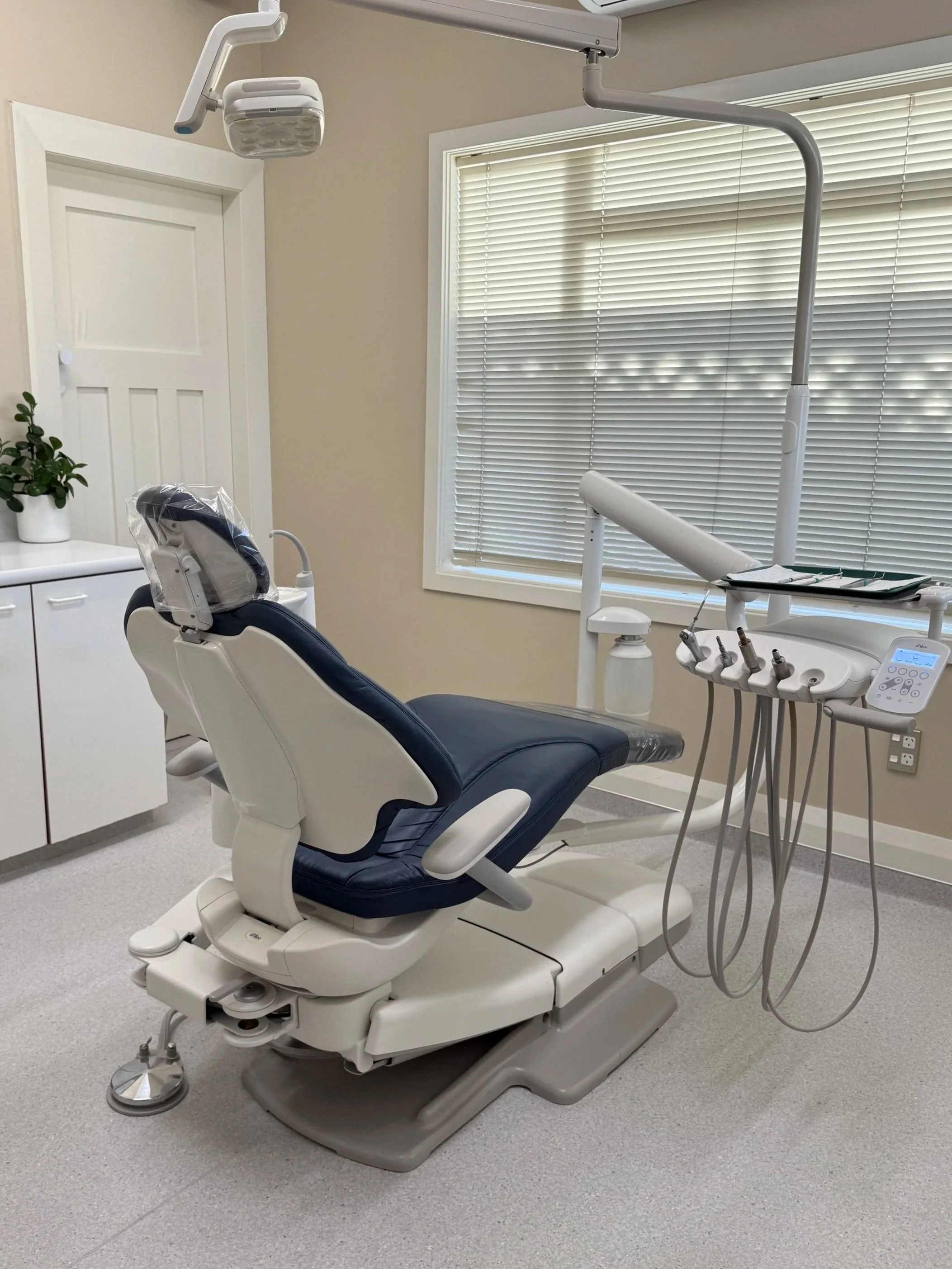 Dental examination room with a dental chair, overhead light, dental instruments, and a window with closed blinds.