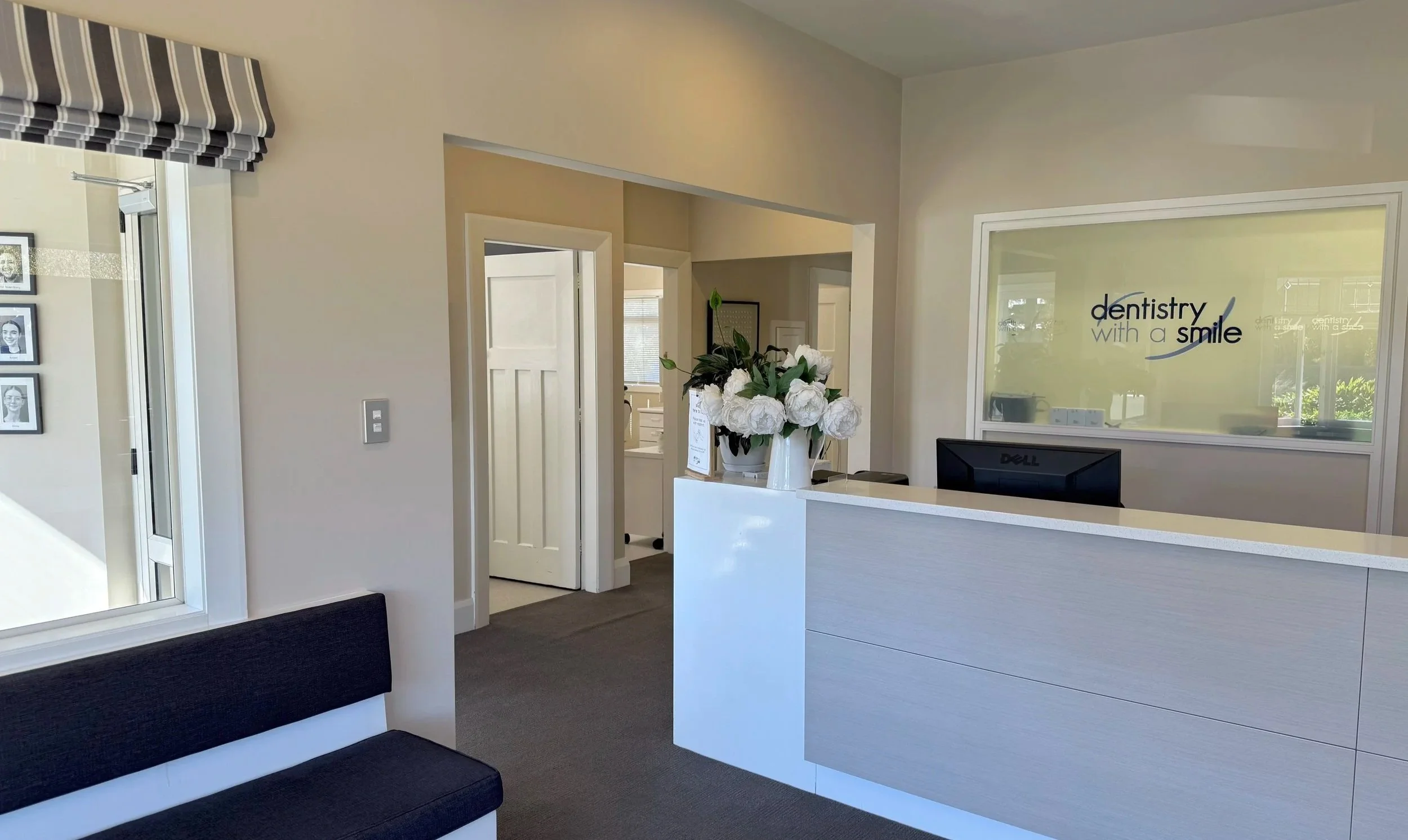 Reception area of a dental office with a white counter, a computer monitor, a large floral arrangement, and a glass window with the words 'dentistry with a smile' printed on it. There is a doorway and a wall with photos on the left.