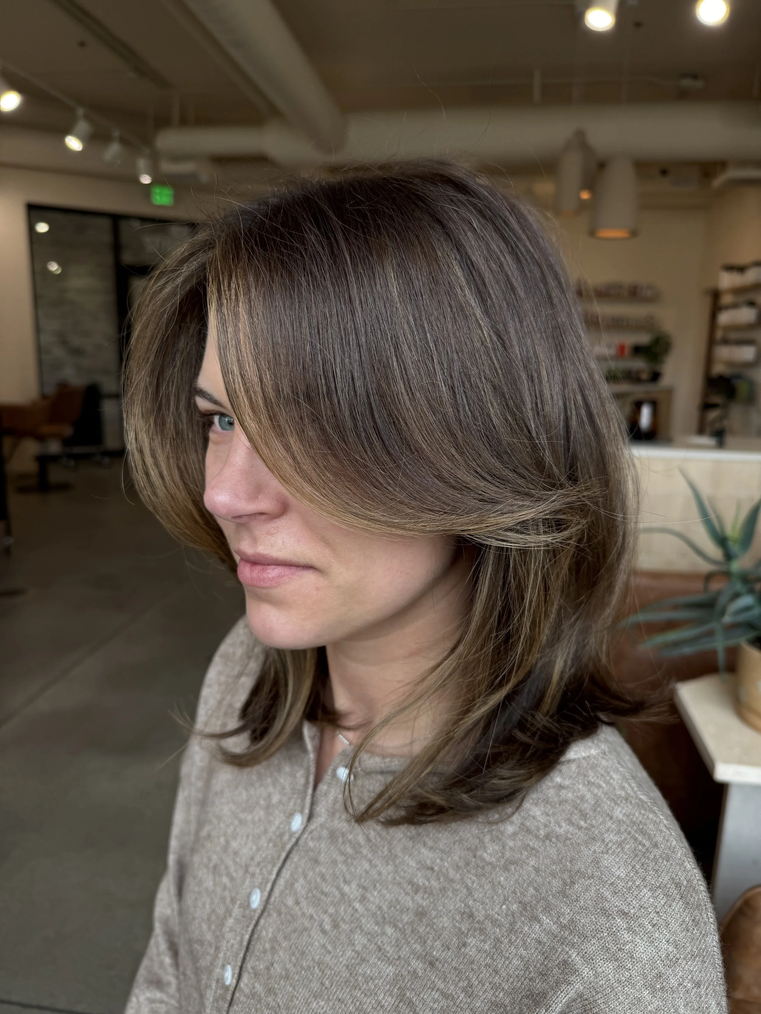 A woman with shoulder-length brown hair styled with soft layers, wearing a beige button-up top, standing in a modern, warmly lit indoor space with bookshelves and plants in the background.