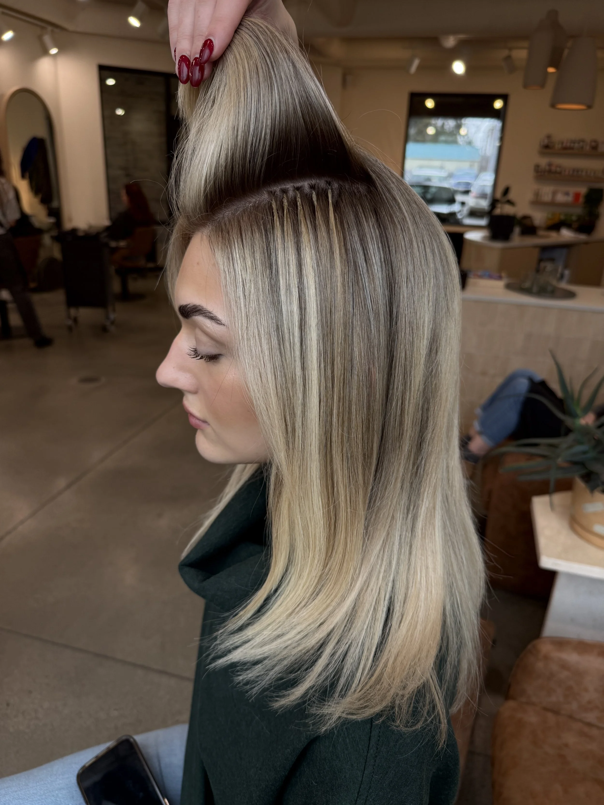 A woman with blonde hair, sitting in a salon, getting her hair styled. A stylist's hand is visible at the top, holding a section of her hair. The salon has large windows, modern decor, and a reception area in the background.