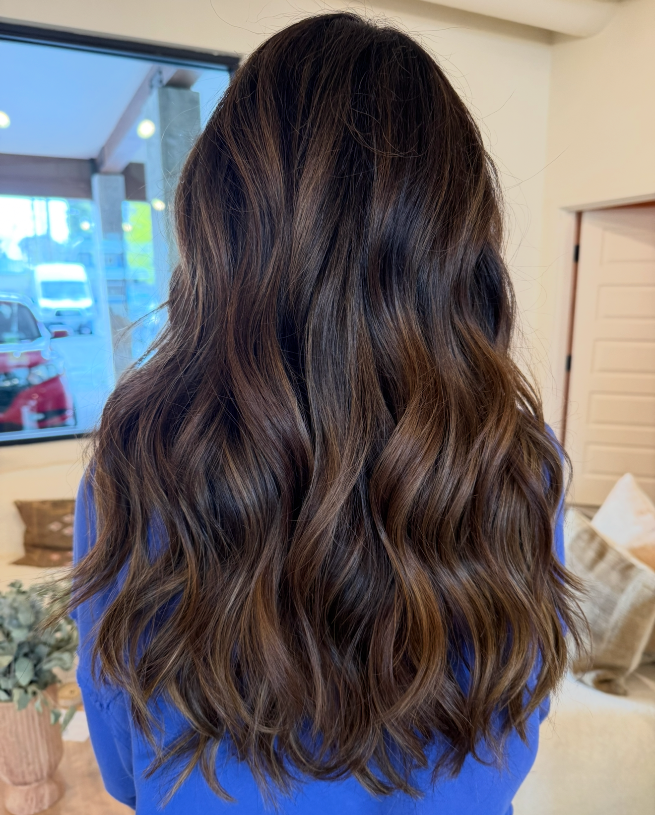 Woman with long, wavy, brunette hair standing indoors, with a window showing a parking lot outside.