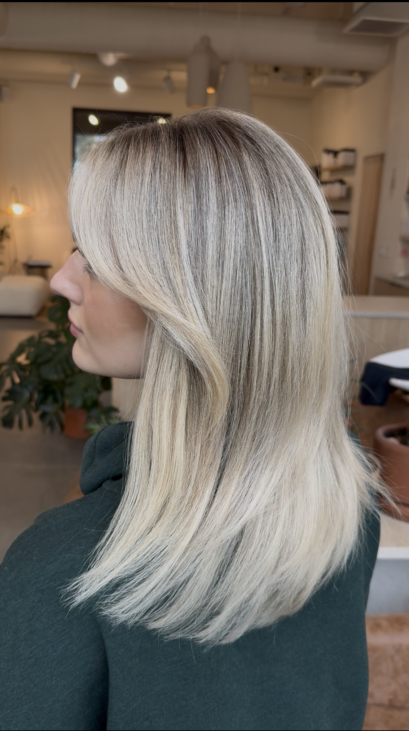 Side view of a woman with long, straight blonde hair, sitting indoors in a well-lit space with a cozy background.