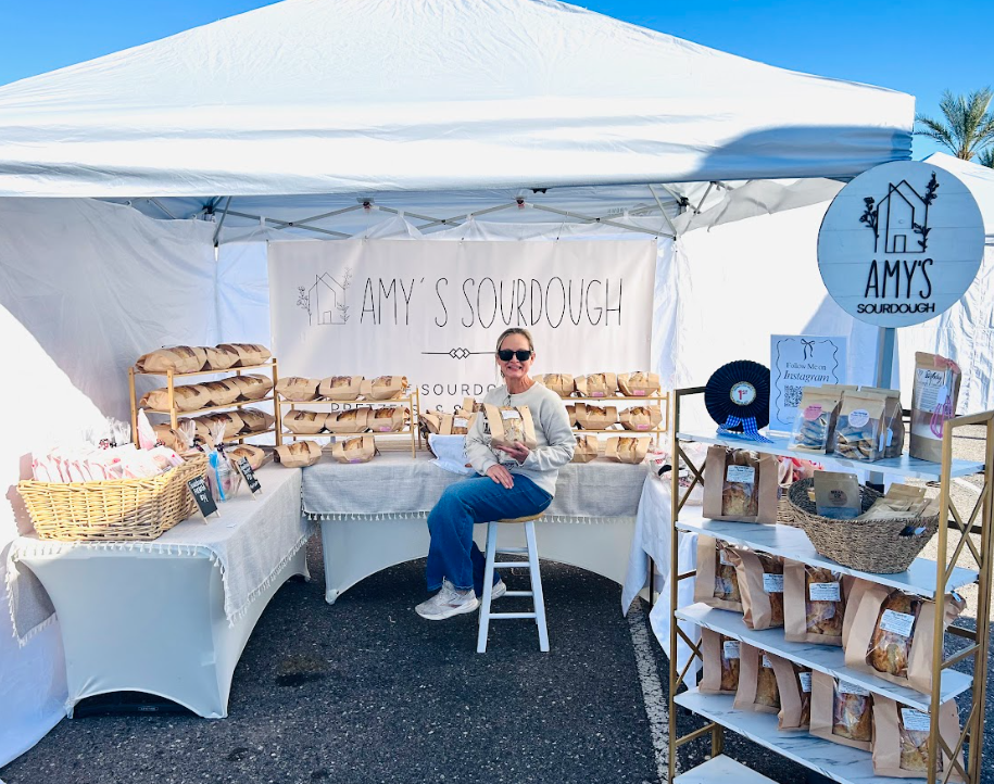 A woman sitting on a stool in front of a stand at an outdoor market selling sourdough bread from Amy's Sourdough, with shelves of bread, a basket of bread, and promotional materials under a white canopy on a sunny day.