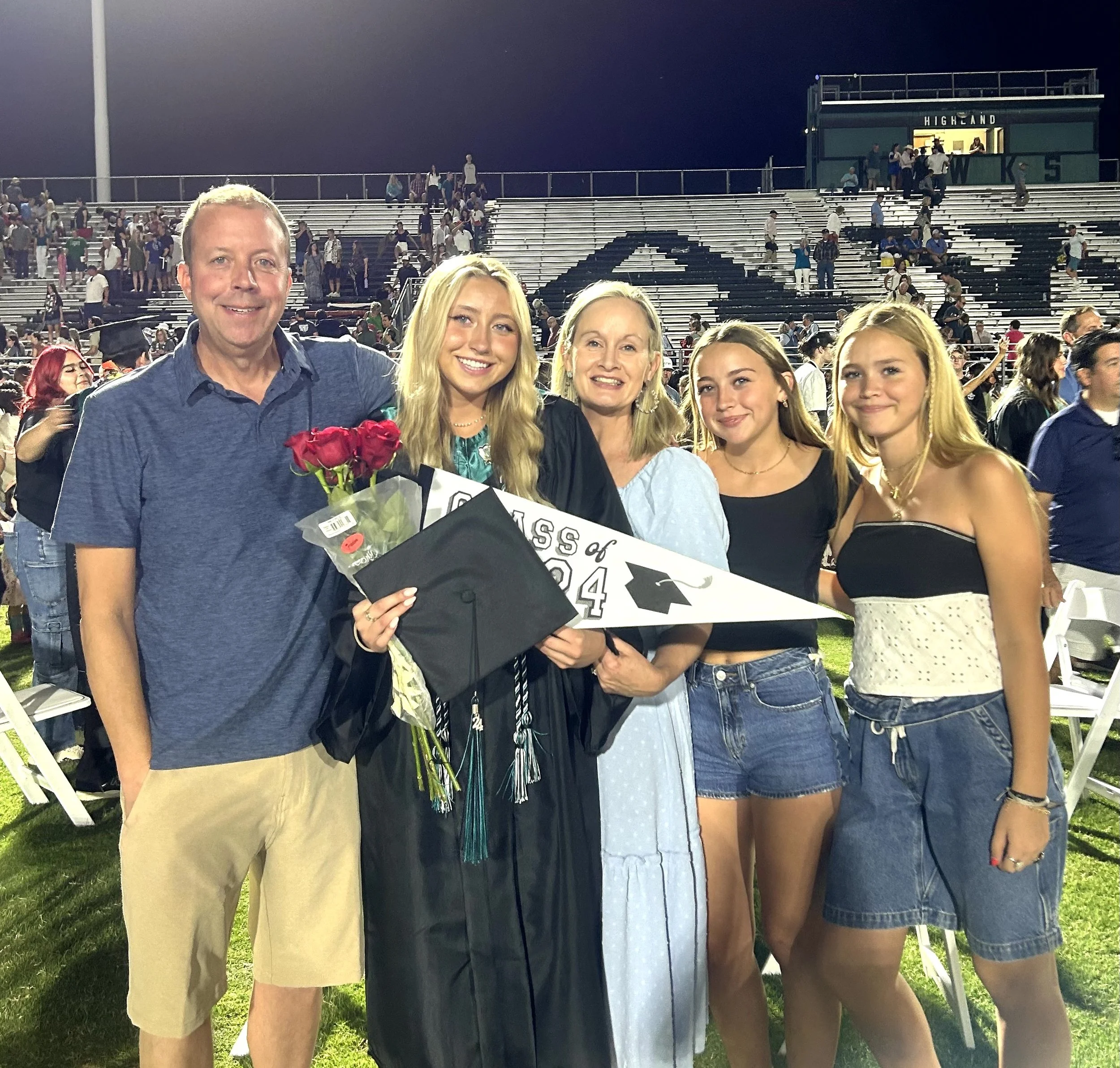 Family celebrating graduation at a stadium with a graduate in cap and gown holding a diploma, flowers, and a class of 2024 pennant, surrounded by friends and family.