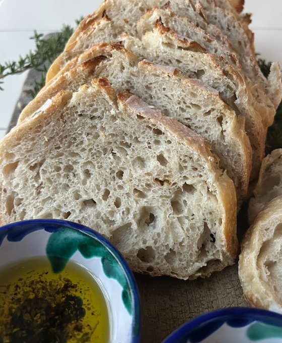 Close-up of sliced rustic bread on a wooden surface with a small bowl of olive oil and herbs nearby.