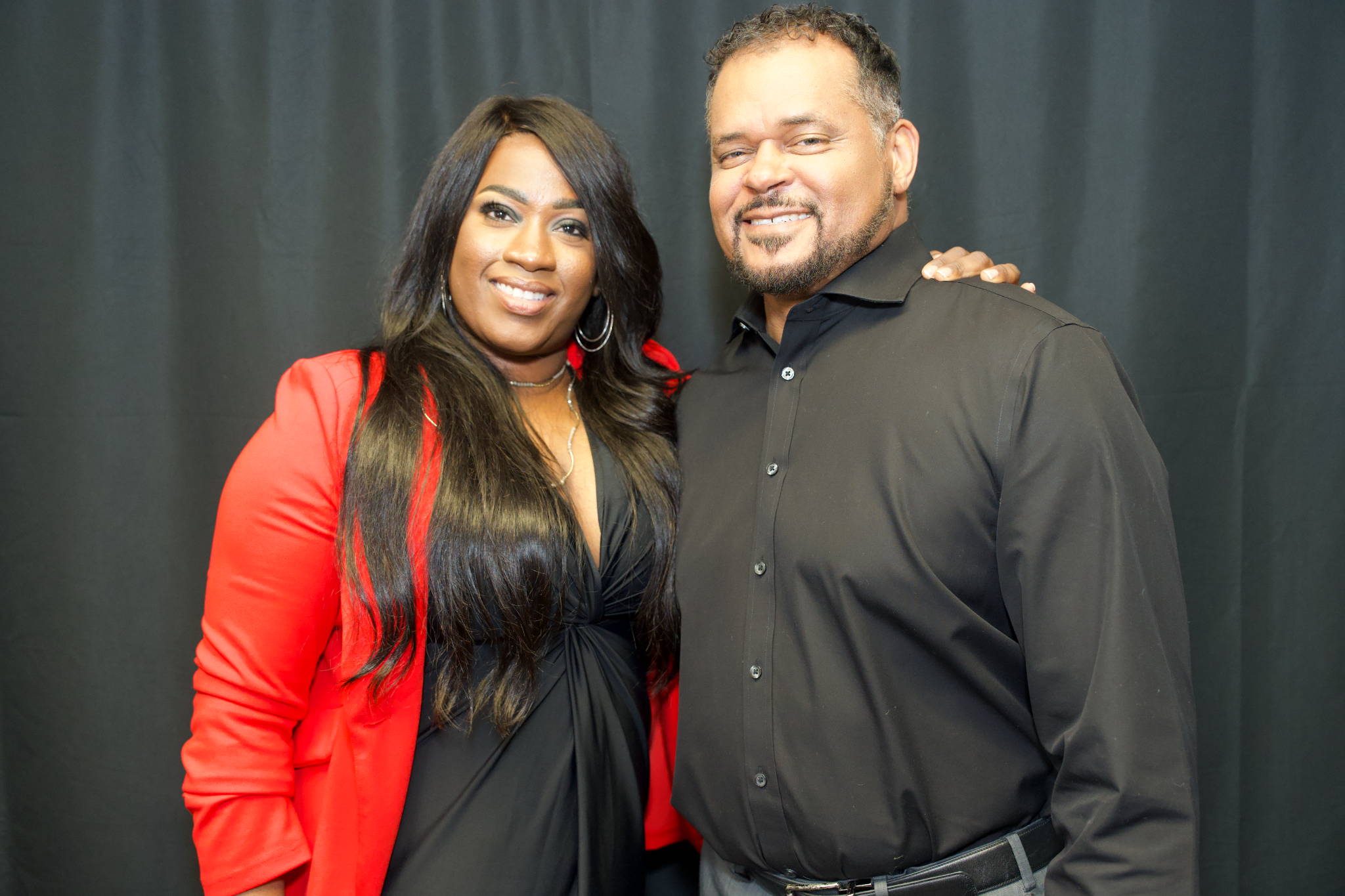 Jacquelyn Clayborn with long dark hair, wearing a red blazer, and Milton Quinn with curly hair, beard, wearing a black shirt, posing together in front of a black background for Say Less Do More Foundation