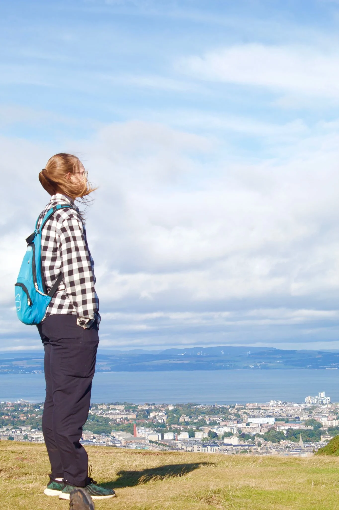 A woman with a backpack standing on a grassy hill overlooking a city, water, and mountains under a blue sky with clouds.
