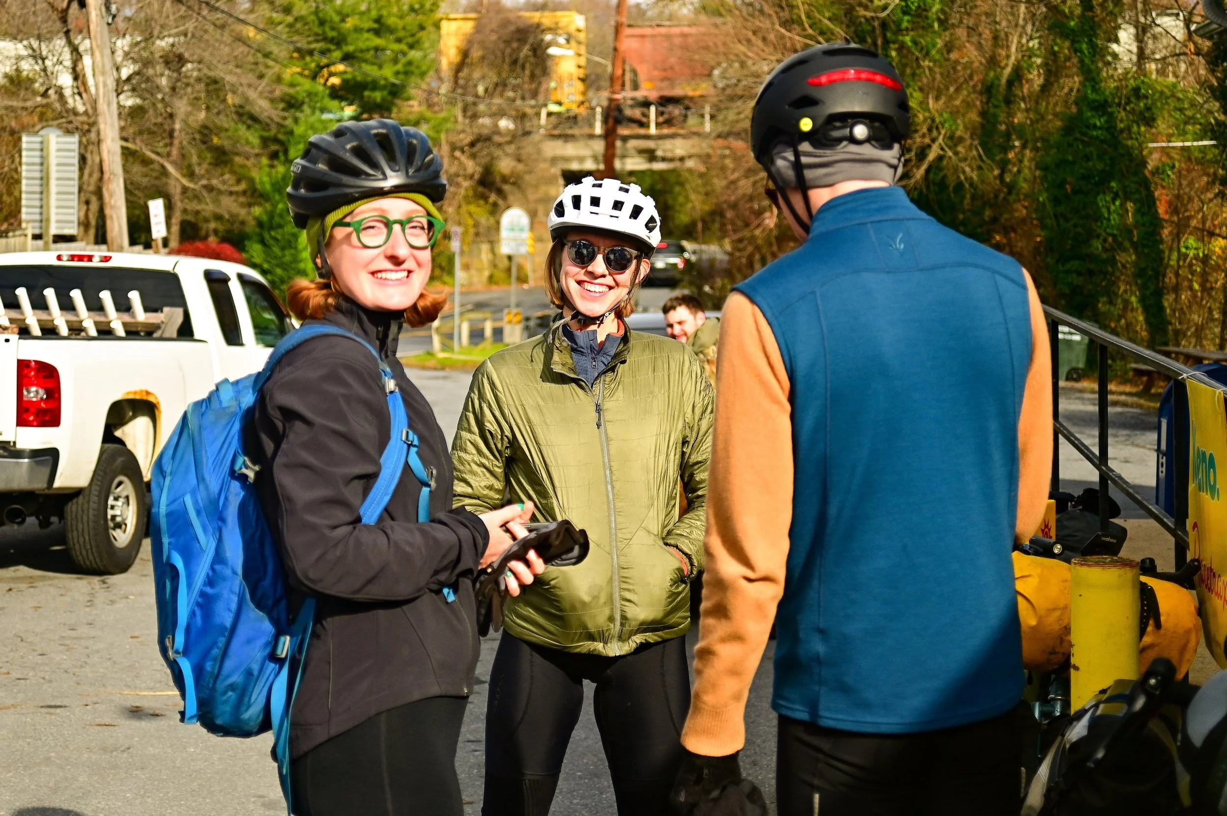 Three cyclists in helmets and outdoor clothing talking and smiling outdoors on a sunny day, with a pickup truck and trees in the background.