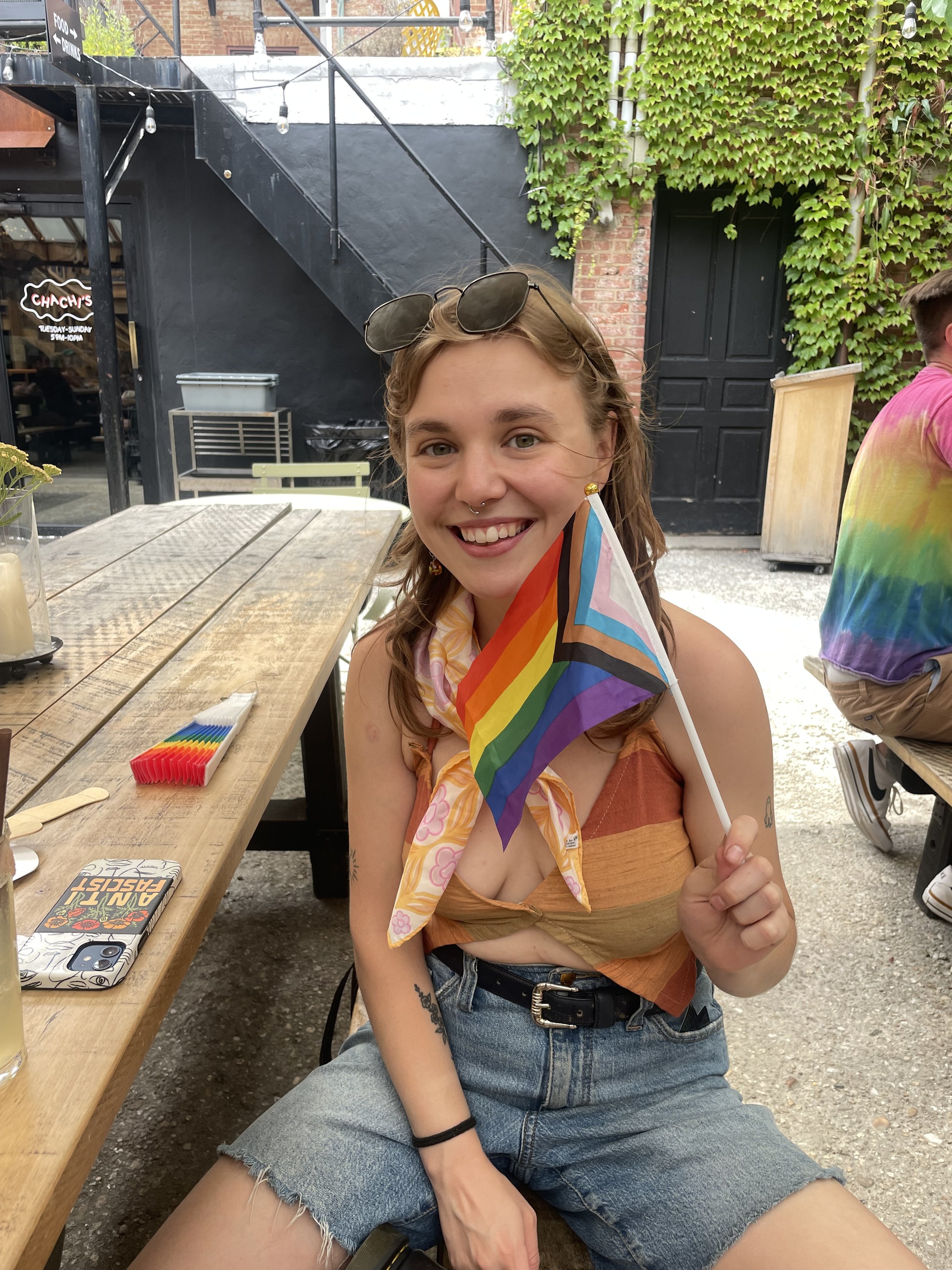 A smiling young woman with glasses on her head and a septum piercing, holding a rainbow pride flag, sitting outdoors at a wooden table with rainbow candles and a phone, wearing a colorful top, denim shorts, and a scarf, celebrating Pride.
